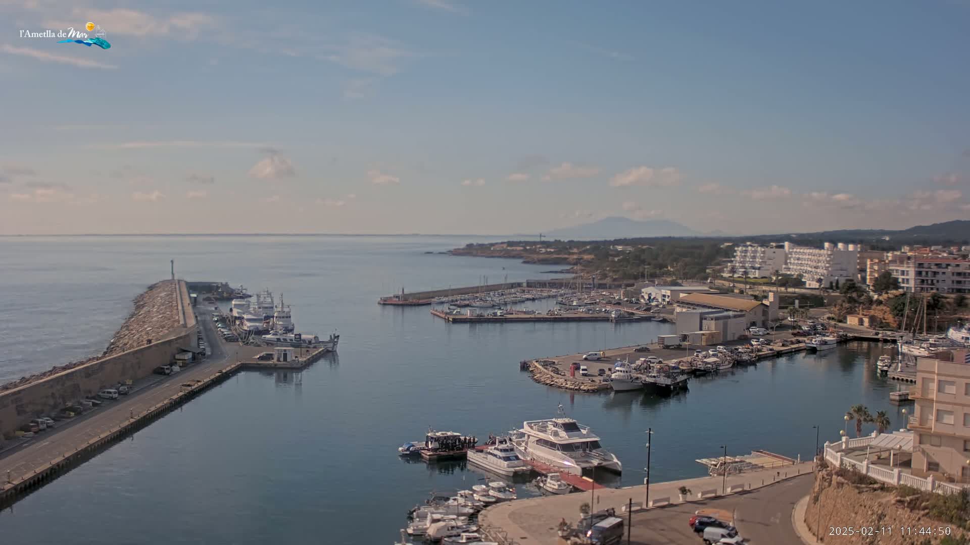 A harbor filled with numerous boats and yachts is situated along a coastline under a partly cloudy sky.