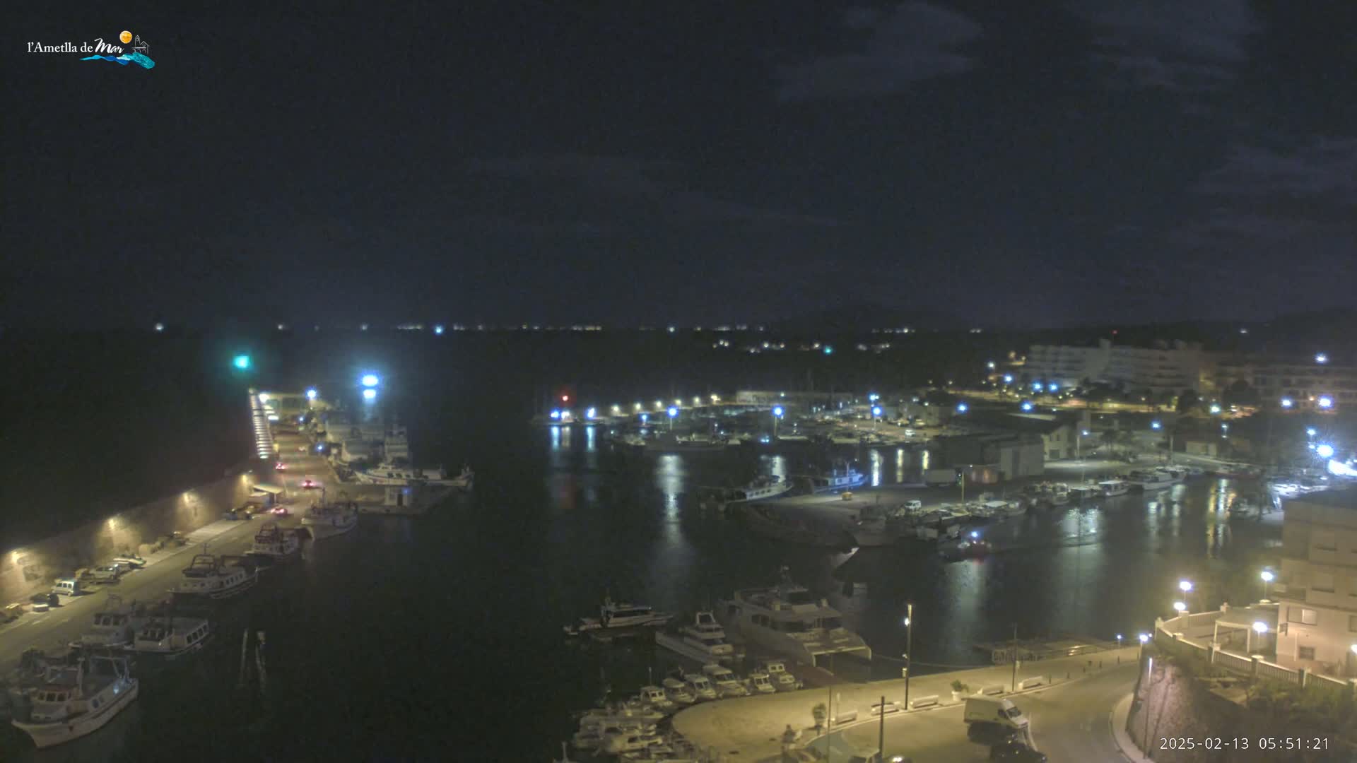 A nighttime aerial view of a harbor filled with boats, illuminated by streetlights and building lights under a partly cloudy sky.