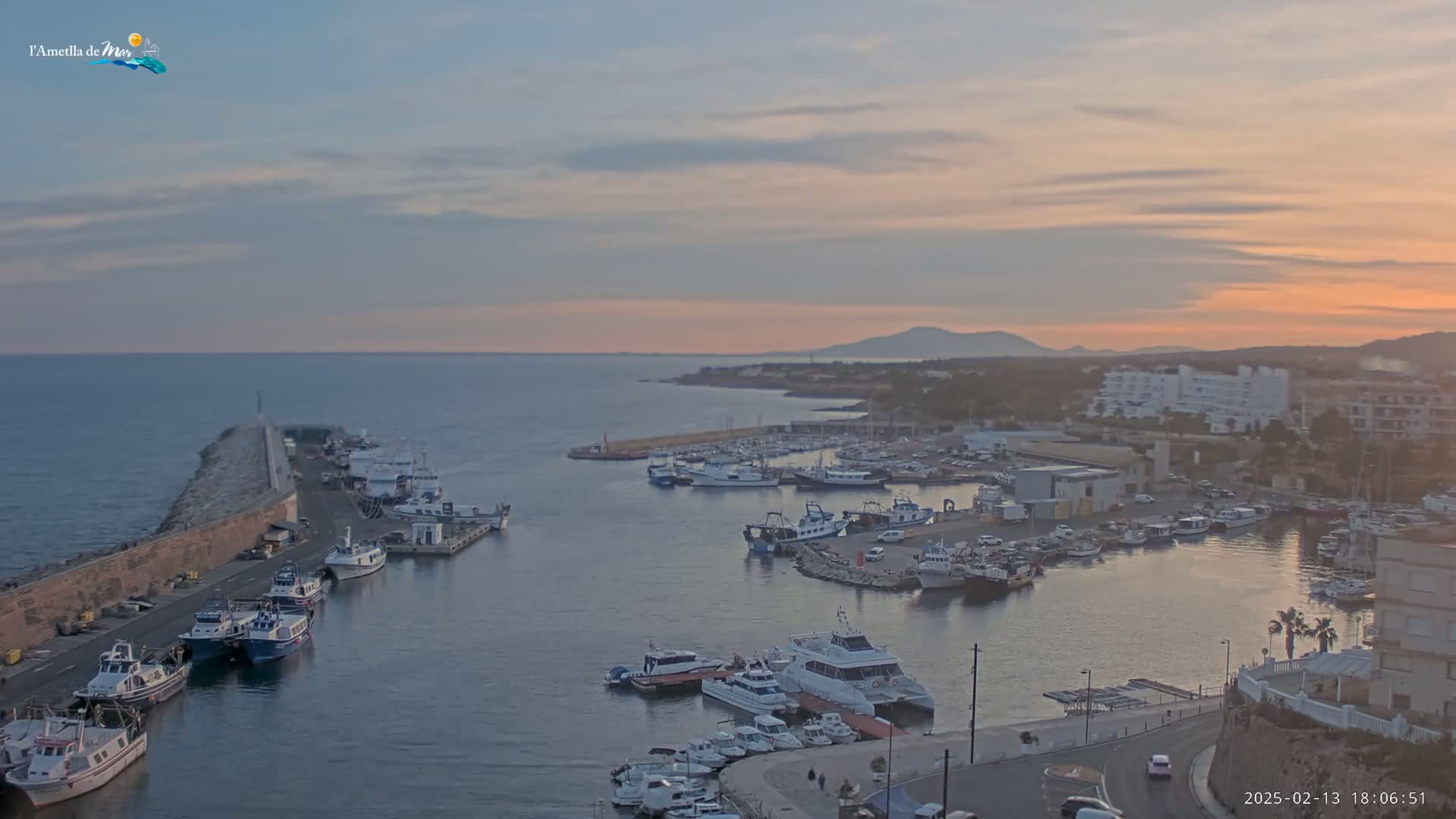 A harbor filled with numerous boats is seen at sunset under a partly cloudy sky, with a coastal town and distant mountains in the background.