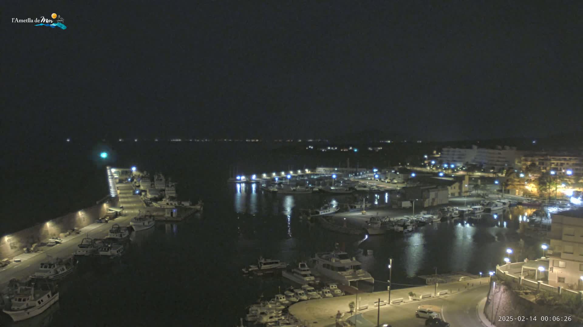 A nighttime, high-angle view of a harbor filled with boats, illuminated by lights, and bordered by buildings and a roadway.