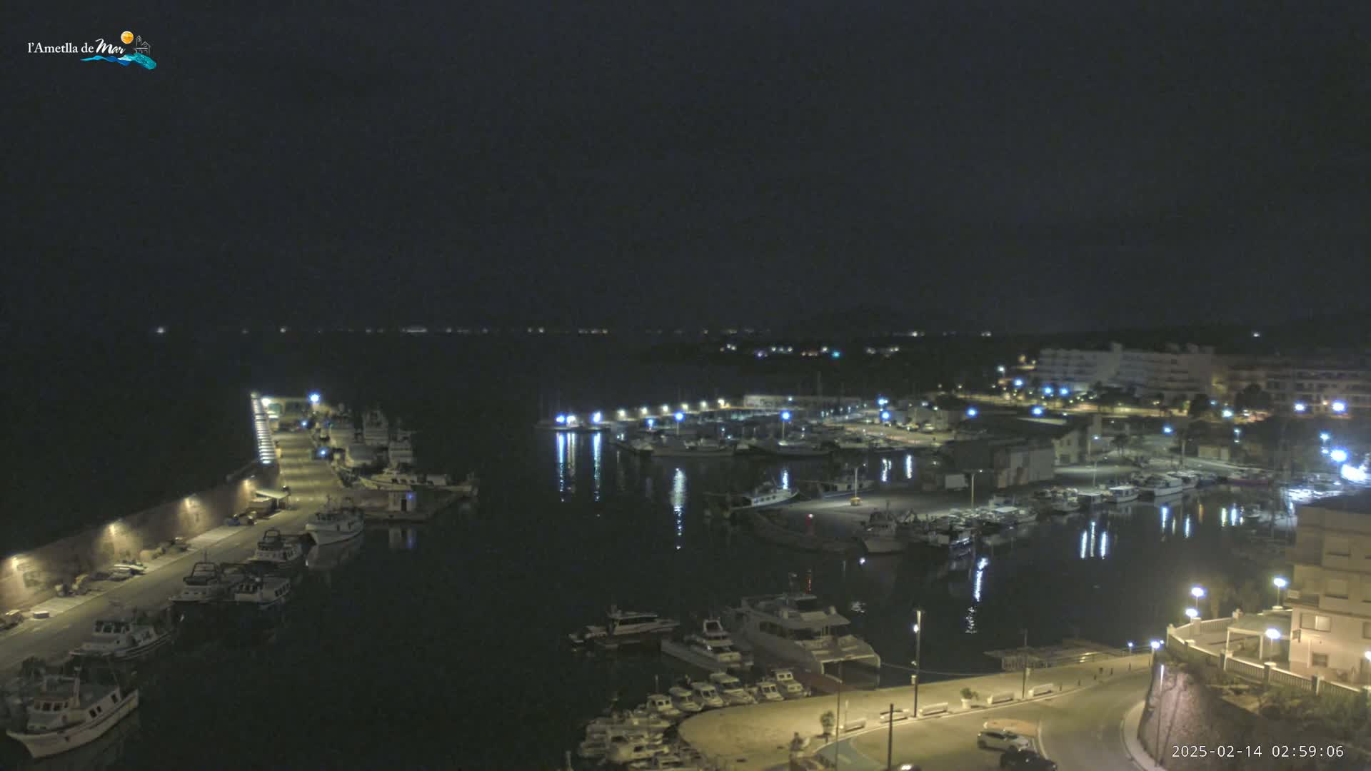 A nighttime view of a harbor filled with boats, illuminated by lights along a waterfront, under a clear, dark sky.