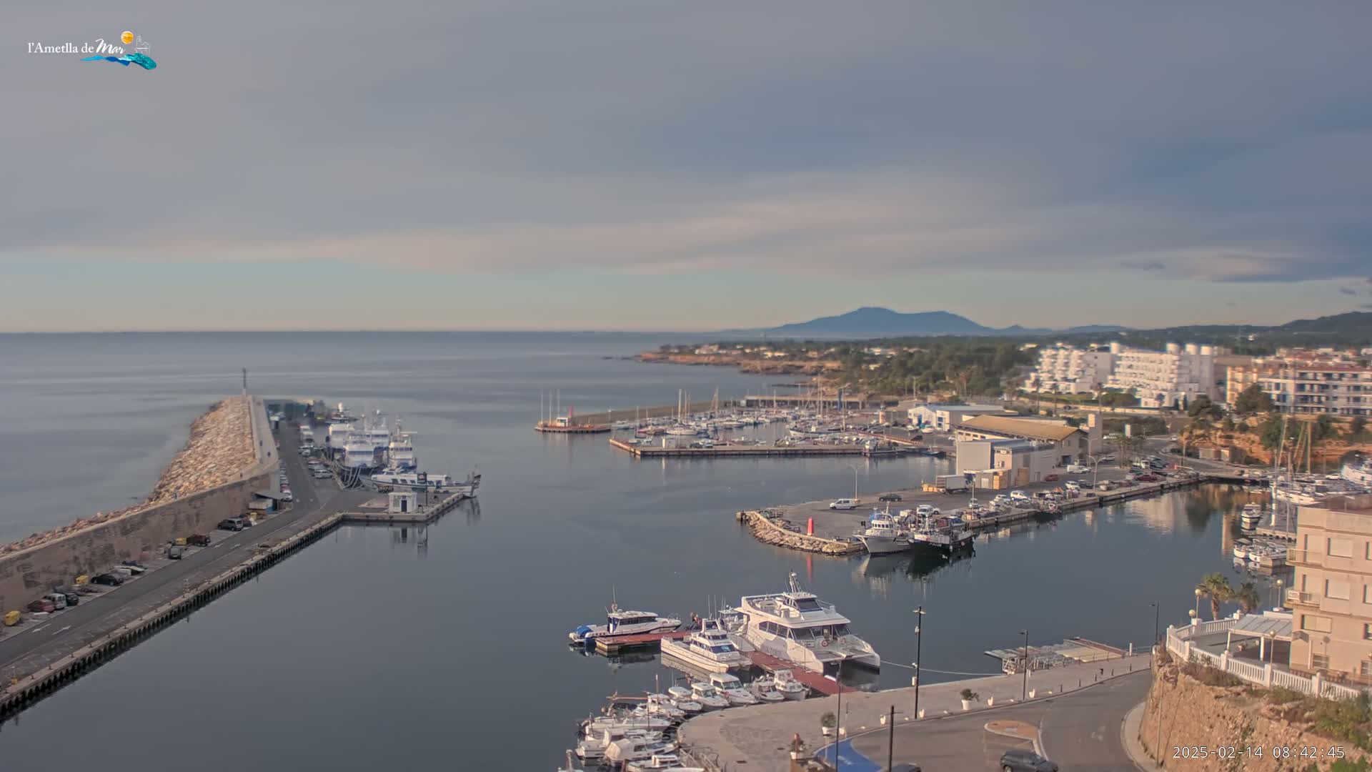 A harbor filled with numerous boats and yachts is situated along a coastline under a partly cloudy sky.