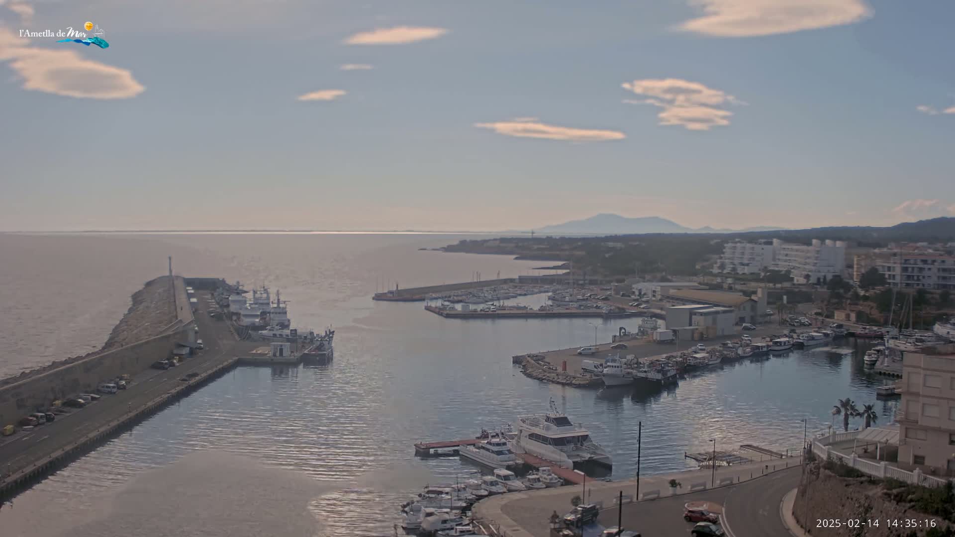 A harbor filled with numerous boats and yachts under a partly cloudy sky, with a calm sea extending to distant mountains.