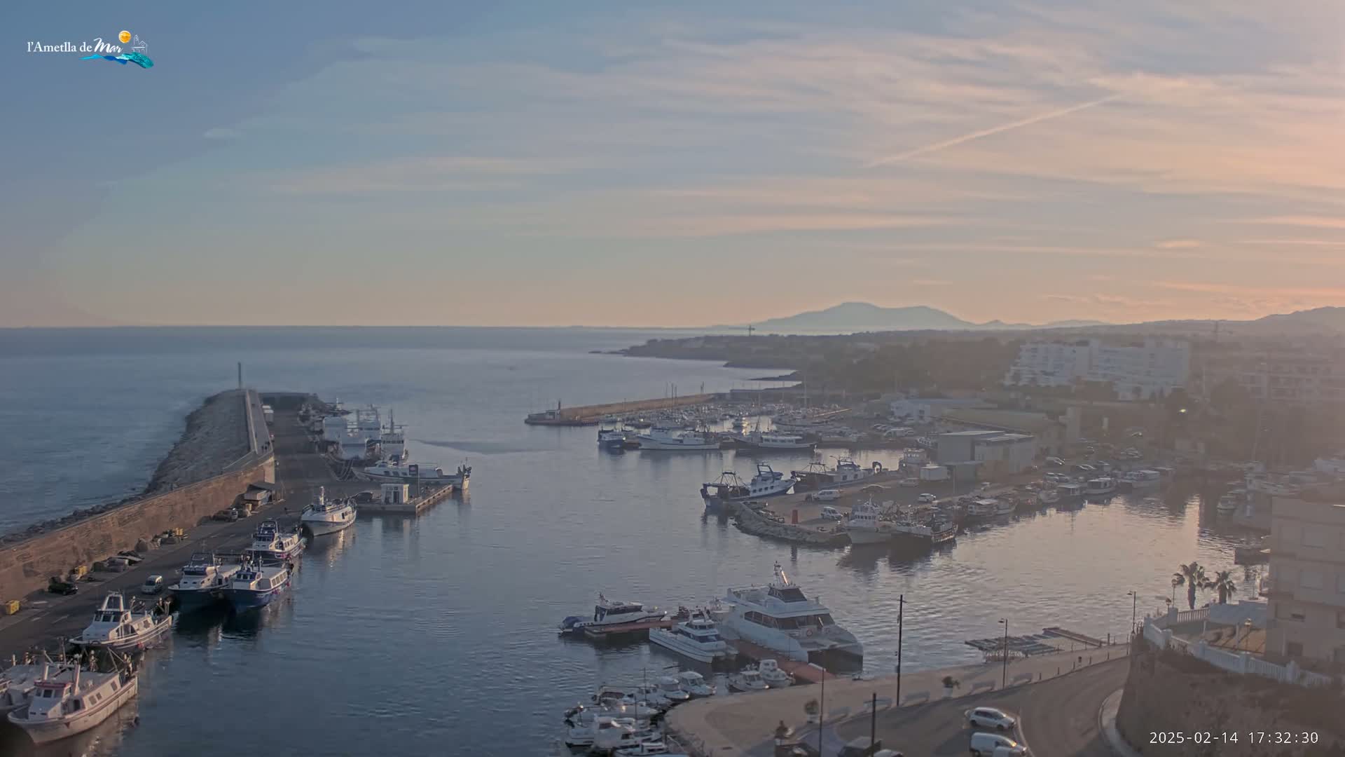 A harbor filled with numerous boats and yachts is shown under a partly cloudy, dusky sky, with a calm sea extending to a distant coastline.