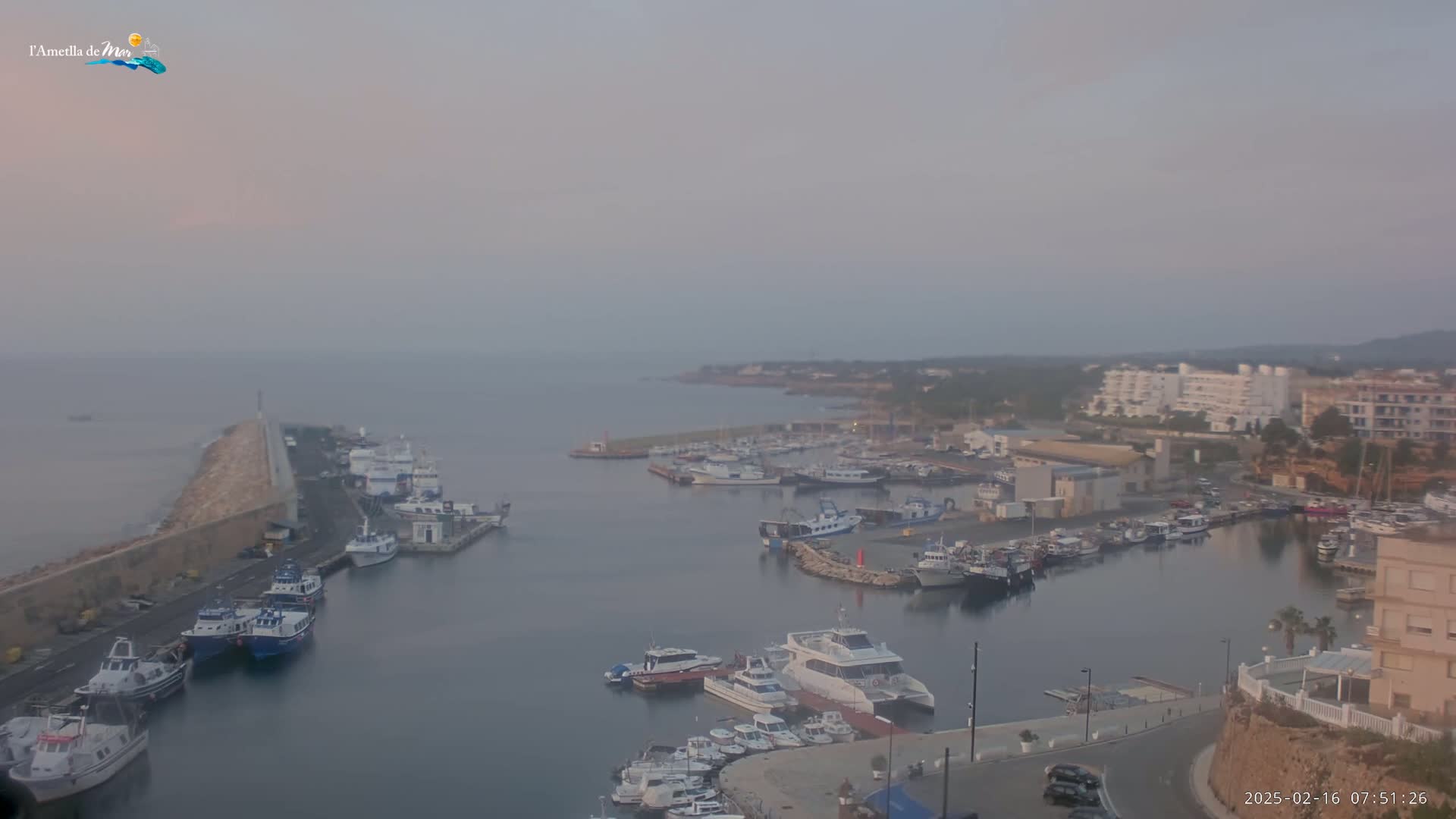 A hazy sunrise illuminates a harbor filled with numerous boats and yachts, with buildings and a coastline visible in the background.