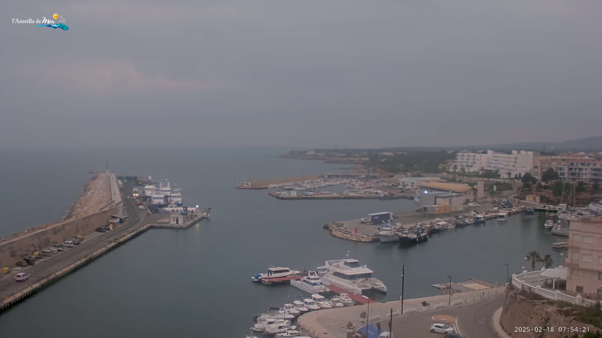 A hazy, overcast day reveals a harbor filled with numerous boats and yachts, alongside a stone breakwater and buildings lining the shore.