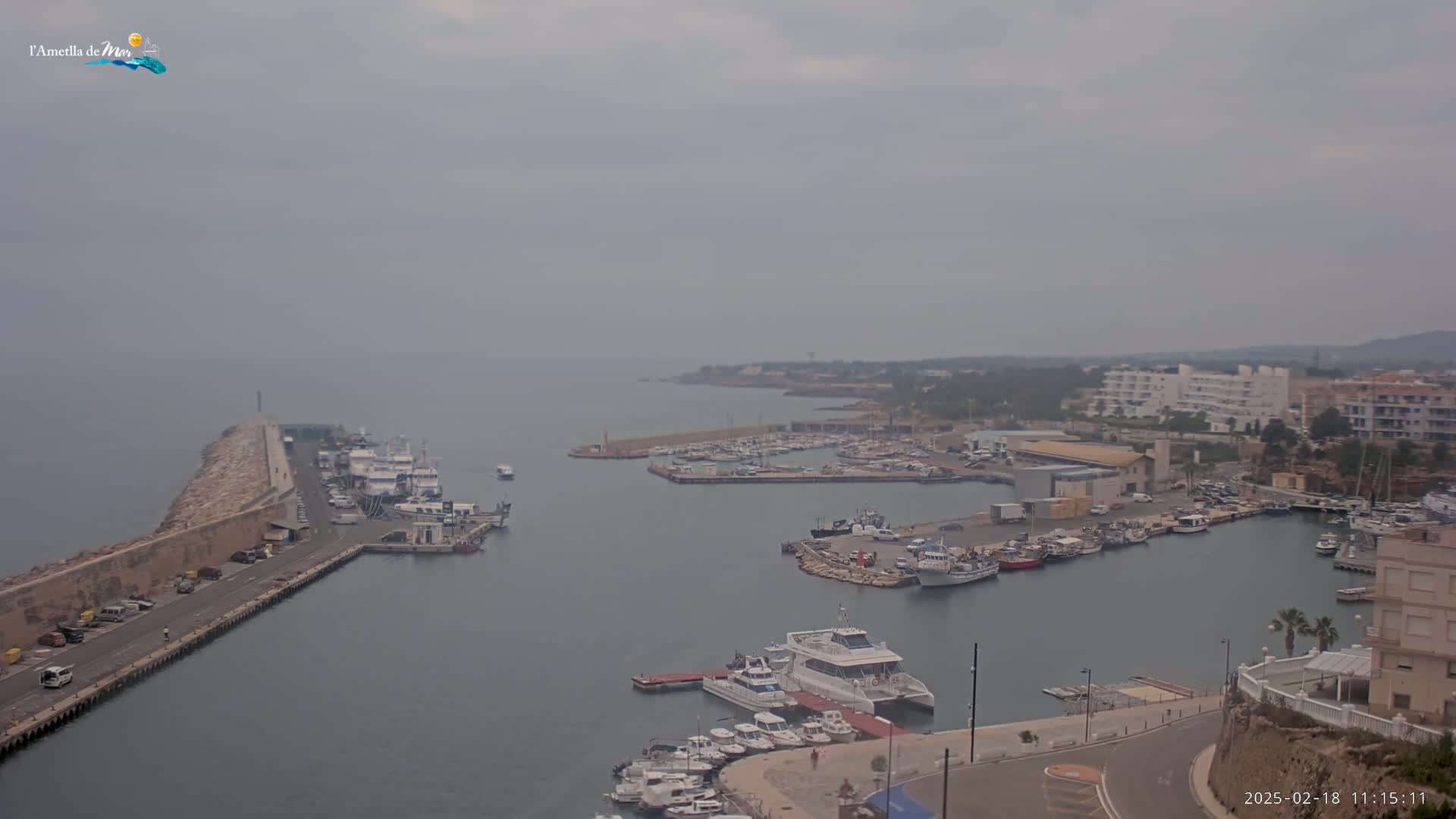 A hazy day reveals a harbor filled with numerous boats and yachts, a long stone breakwater, and buildings along the shoreline.