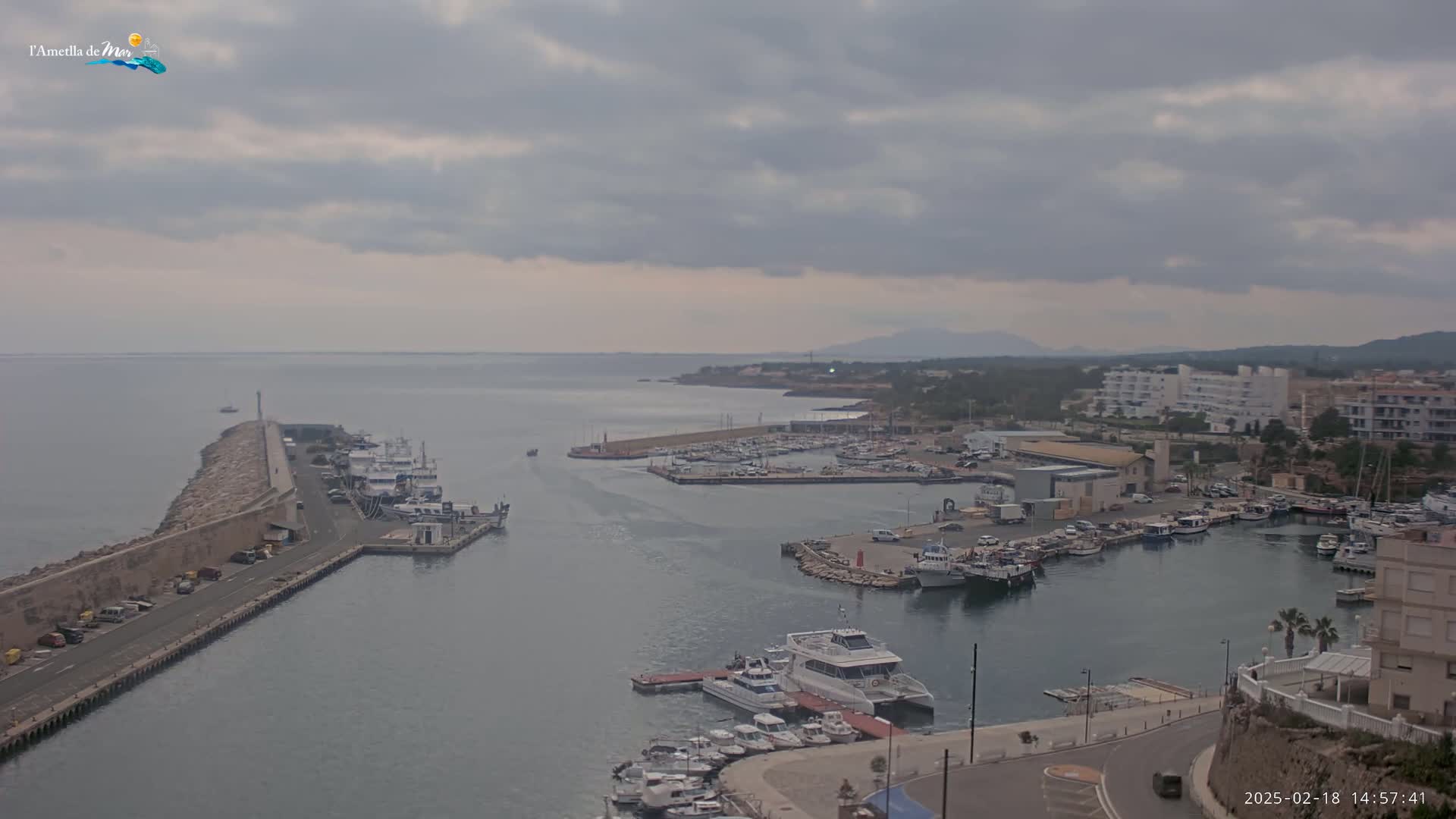 A harbor filled with boats is seen under a cloudy sky, with a road and buildings visible along the shoreline.