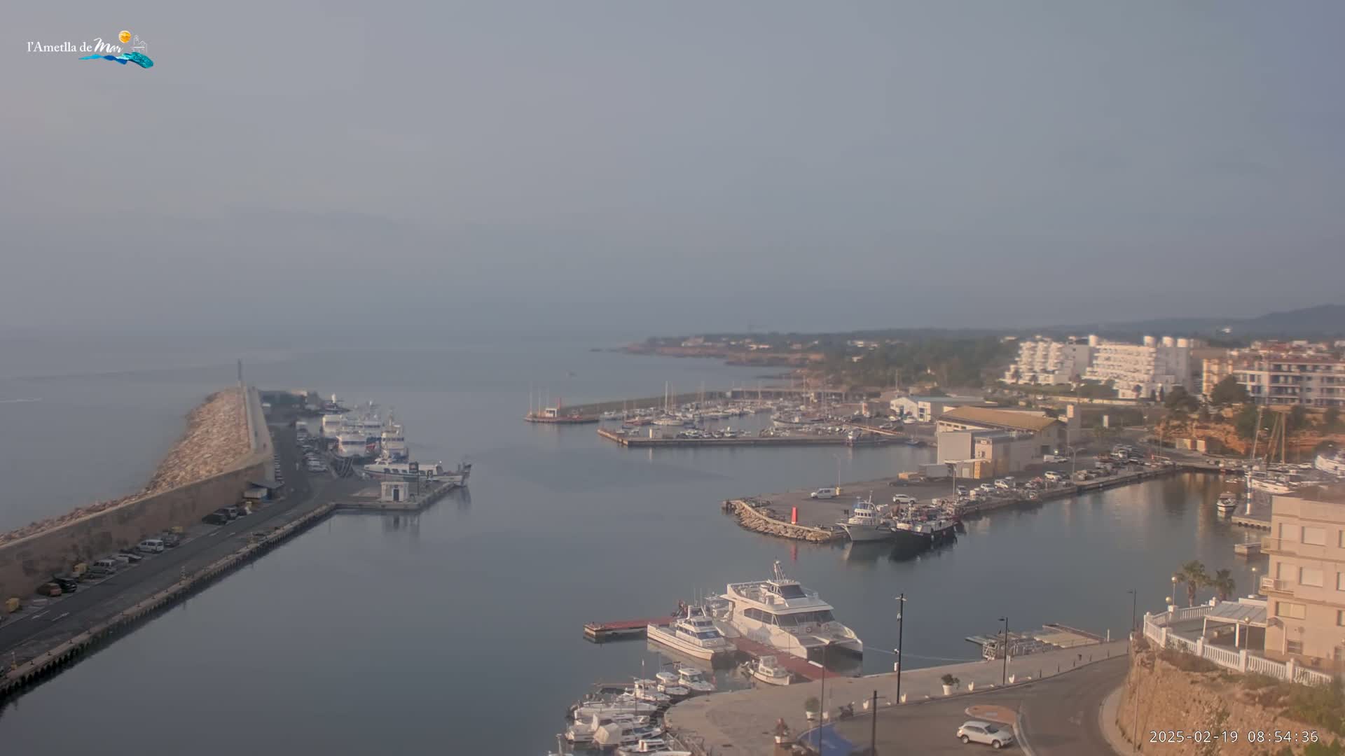 A hazy, calm day reveals a marina filled with boats, a long breakwater, and buildings along a coastline.