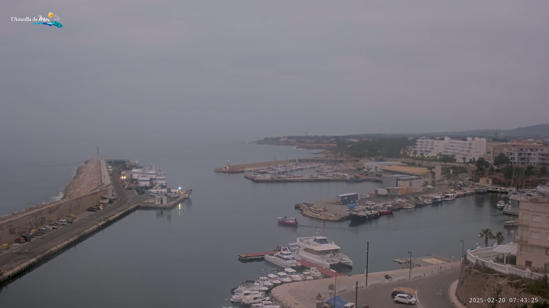 A hazy, overcast morning reveals a harbor filled with numerous boats, a breakwater, and buildings along the coast.