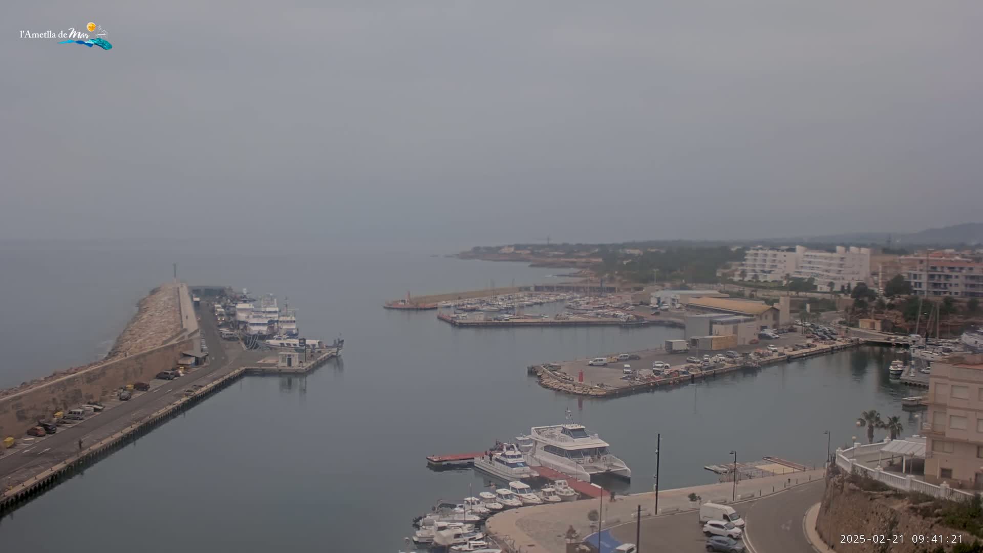 A hazy harbor filled with numerous boats and yachts is seen from an elevated vantage point, with a stone breakwater extending into the calm water.
