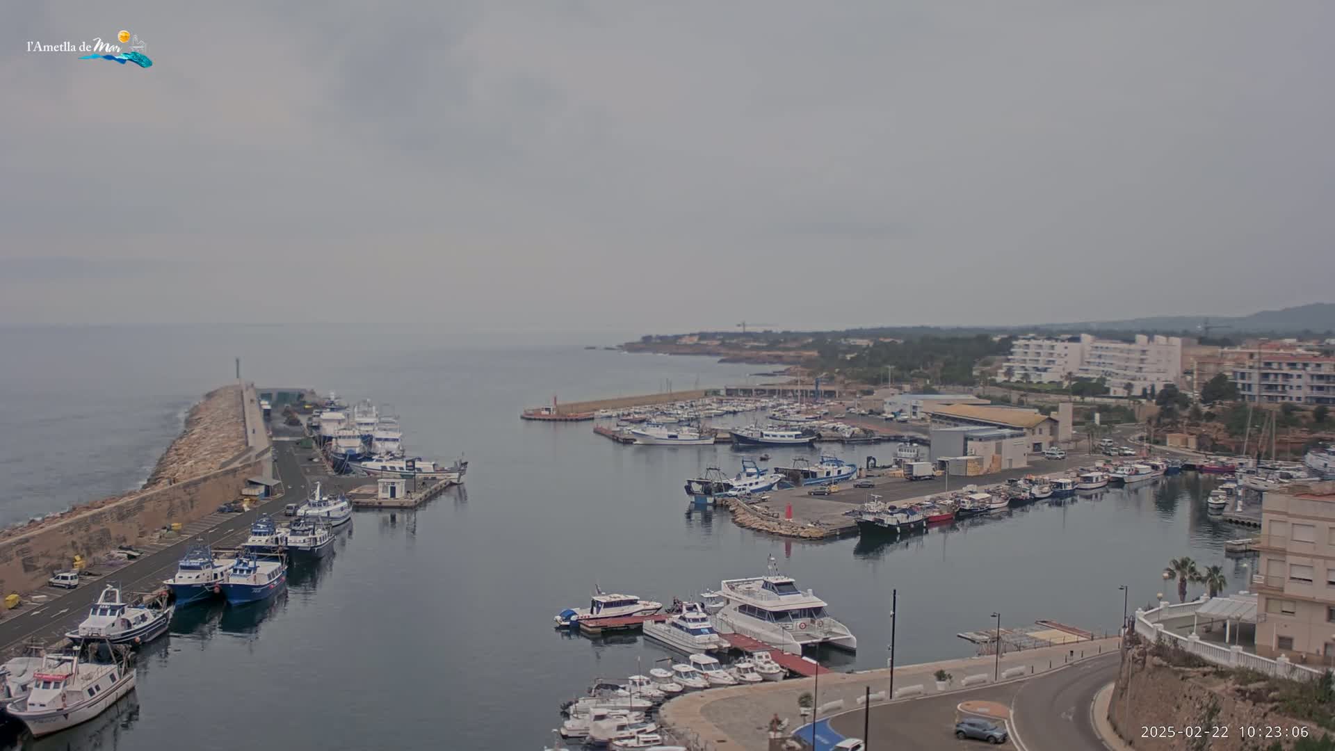 A harbor filled with numerous boats and yachts is seen on an overcast day, with a stone breakwater extending into the calm sea.