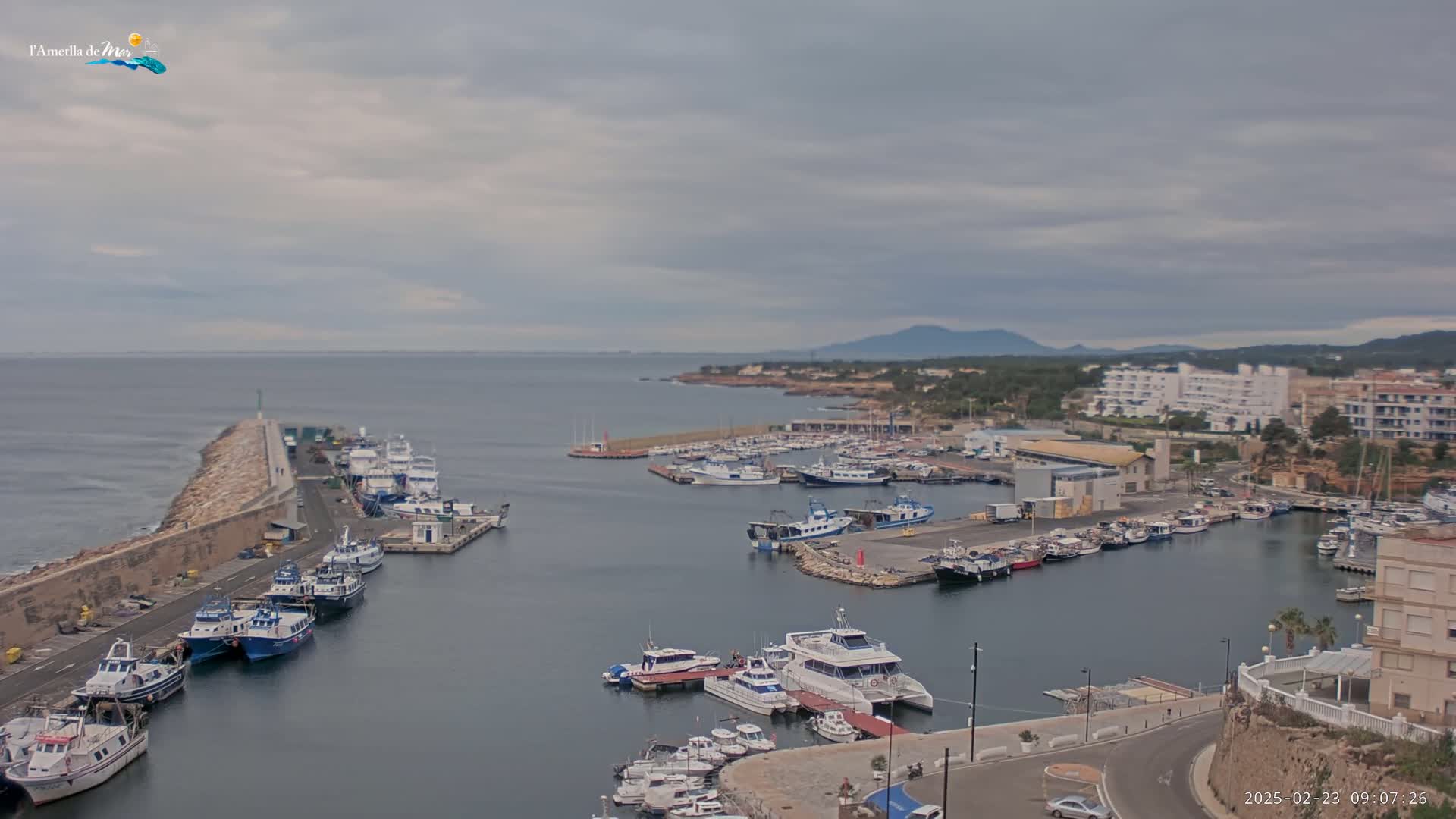 A harbor filled with numerous boats and yachts is shown under an overcast sky, with a calm sea and distant hills visible in the background.
