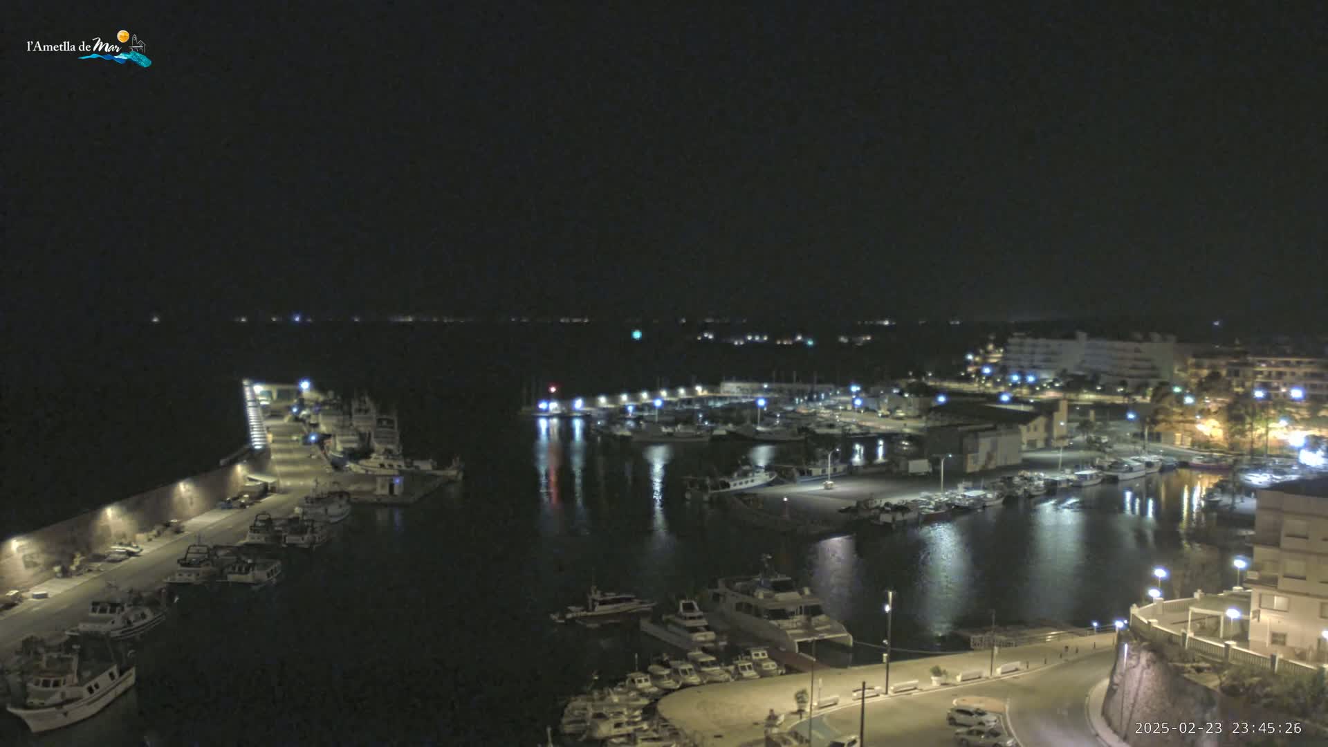 A nighttime aerial view of a harbor filled with boats, illuminated by lights along a paved waterfront, under a clear, dark sky.