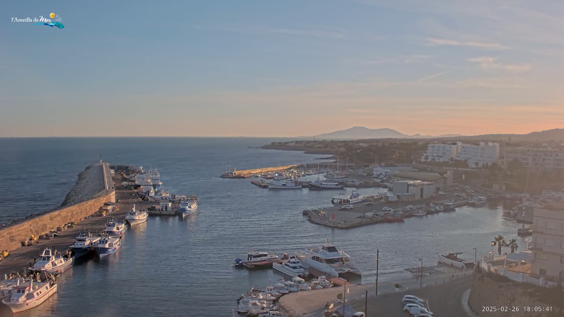 A harbor filled with numerous boats and ships is situated along a coastline under a clear, sunset sky.