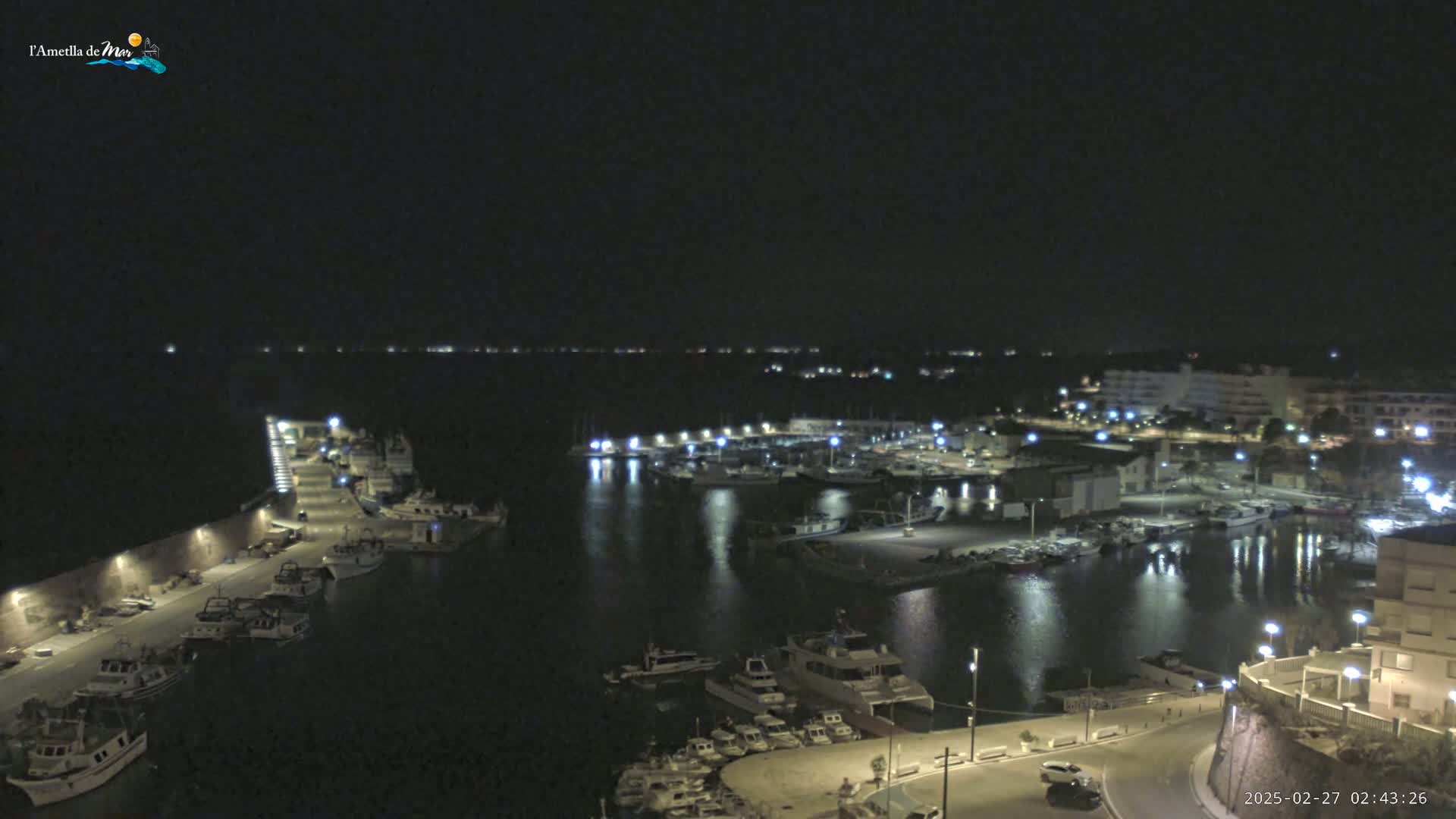 A nighttime view of a harbor filled with boats, illuminated by lights along the docks and buildings on shore under a clear, dark sky.