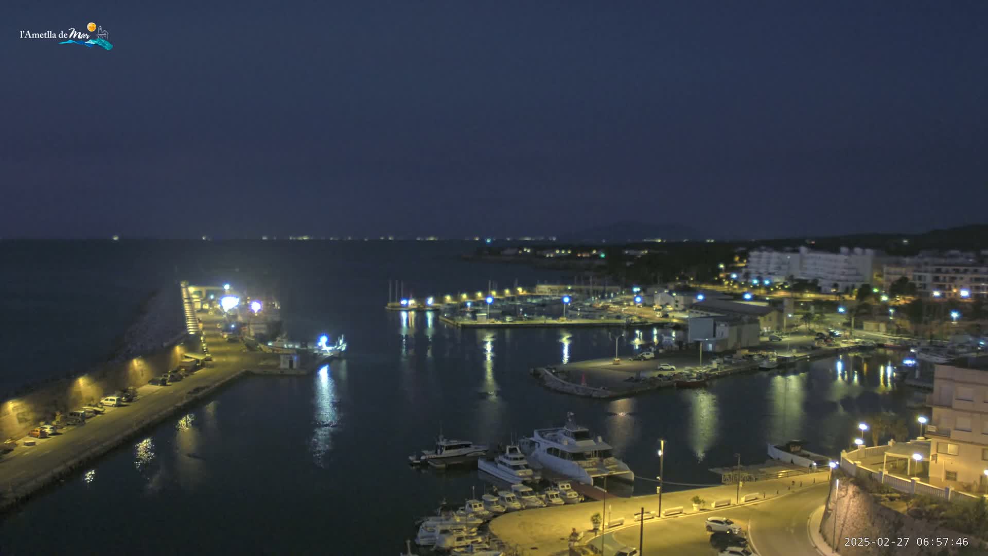 A nighttime harbor scene shows a marina filled with boats, a town with illuminated buildings, and a calm, dark body of water extending to a distant shoreline under a dark sky.