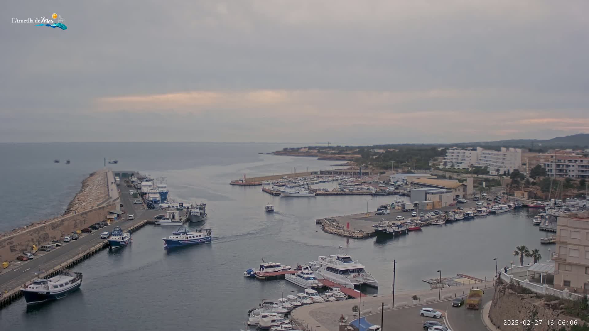 A harbor filled with numerous boats and yachts is seen on an overcast day, with calm water and a stone breakwater extending into the sea.