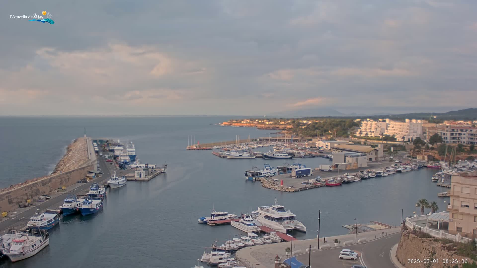 A harbor filled with numerous boats and yachts is situated along a coastline under a mostly cloudy sky.