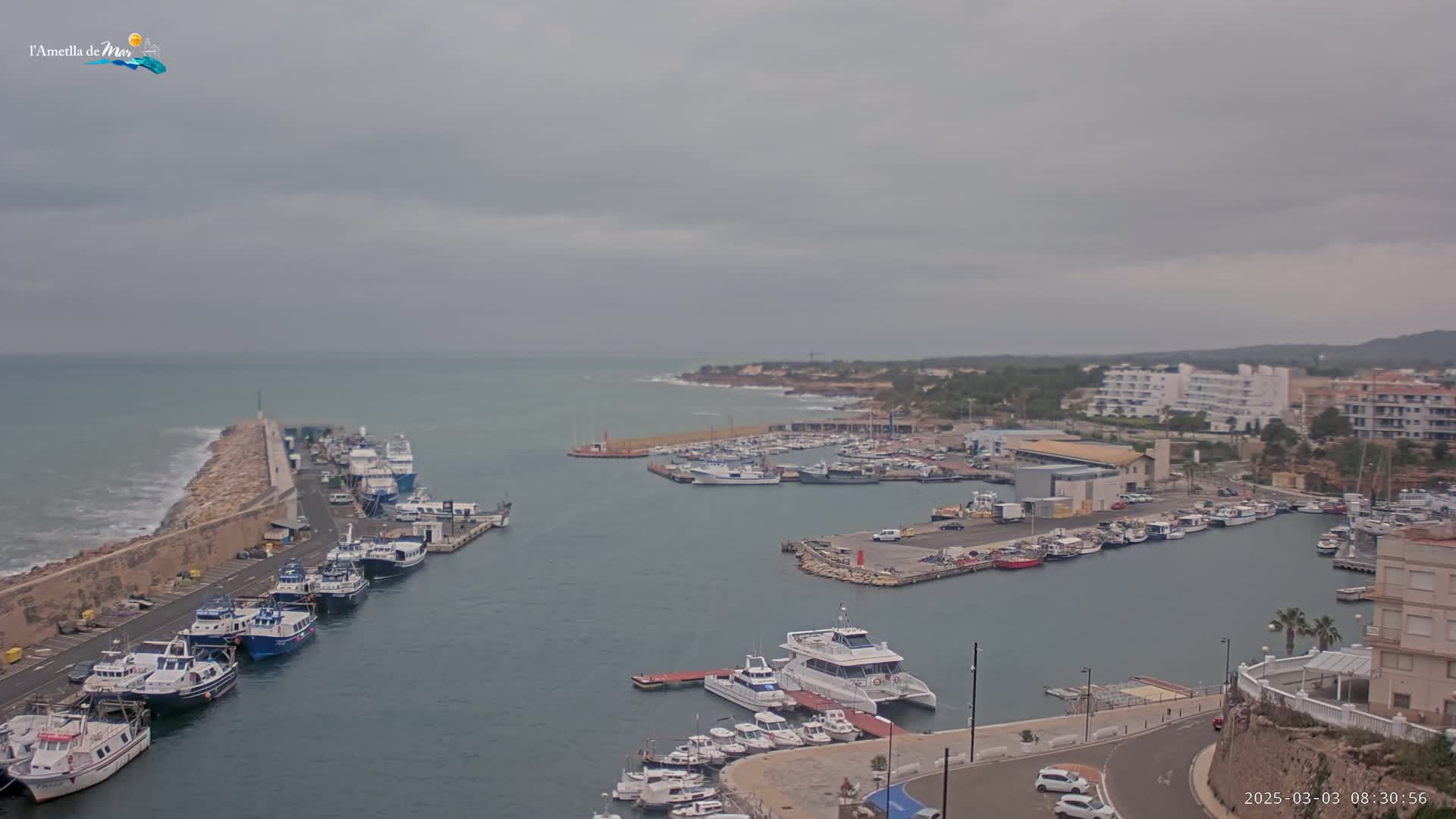 A harbor filled with numerous boats and ships is seen under an overcast sky, with waves breaking against a stone barrier on the left.