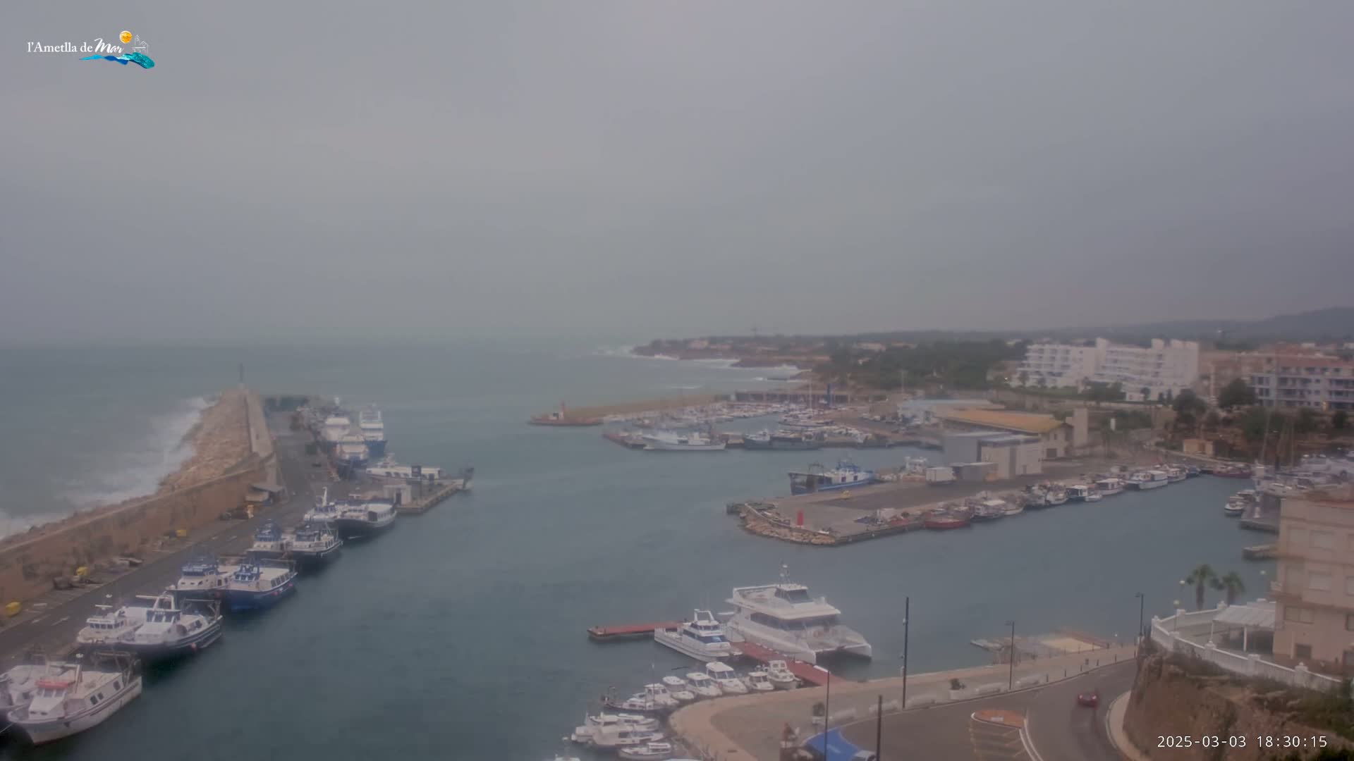 A harbor filled with numerous boats is shown under an overcast sky, with waves crashing against a breakwater.