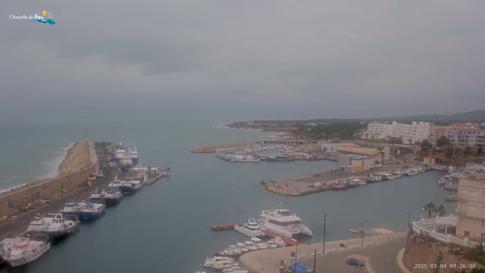 A harbor filled with numerous boats and ships is seen on an overcast day, with a stone breakwater extending into the sea.