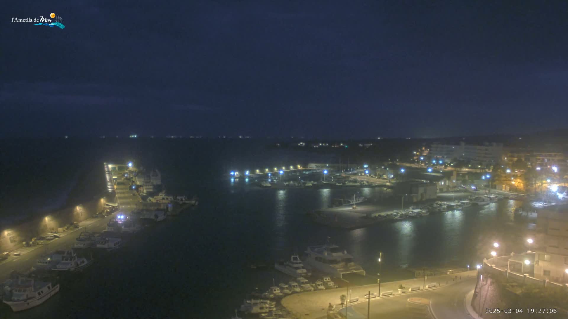 A nighttime view of a harbor filled with boats, illuminated by lights along the waterfront under a dark, clear sky.
