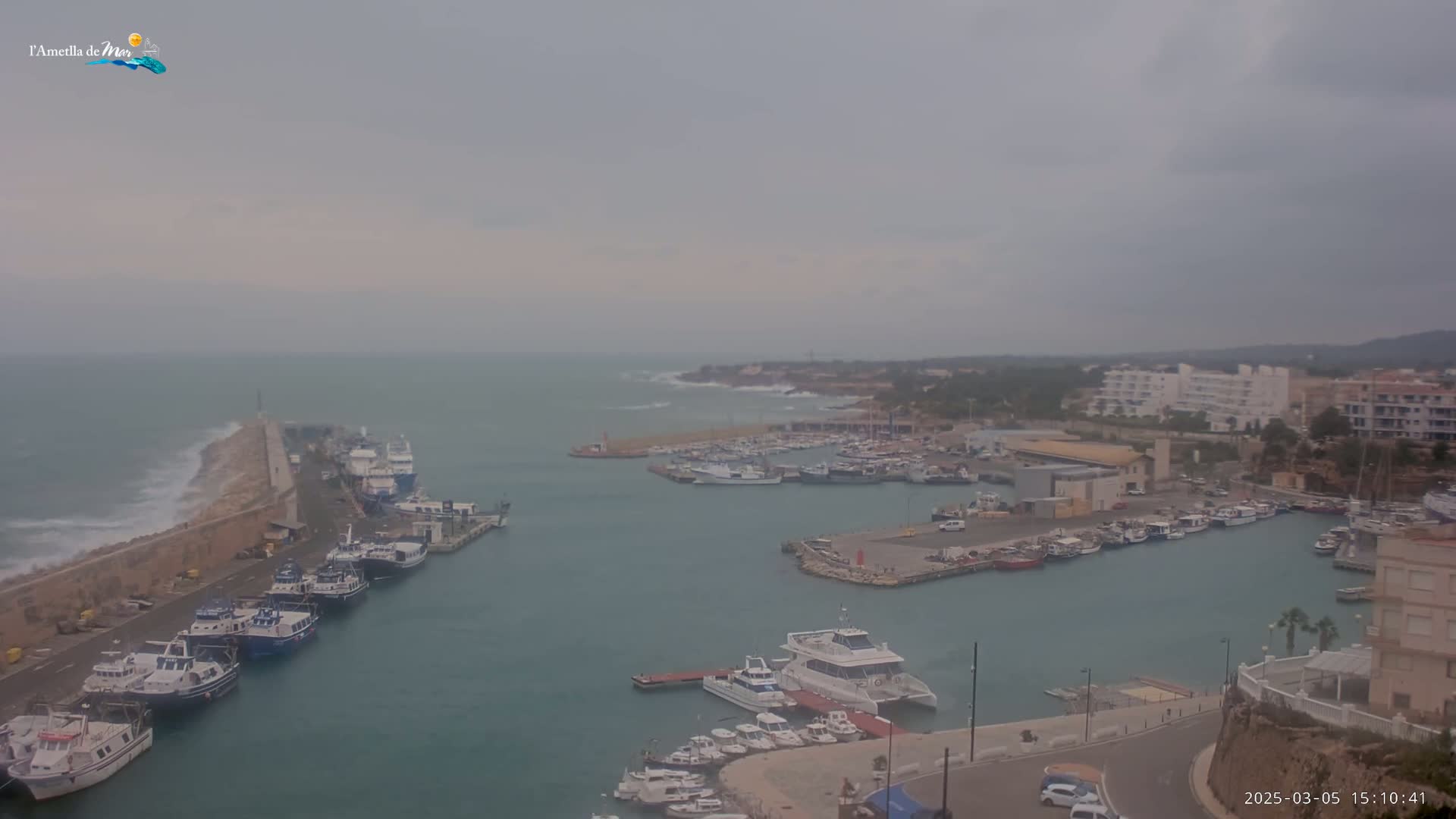 A harbor filled with numerous boats is seen under an overcast sky, with waves crashing against a breakwater in the foreground.