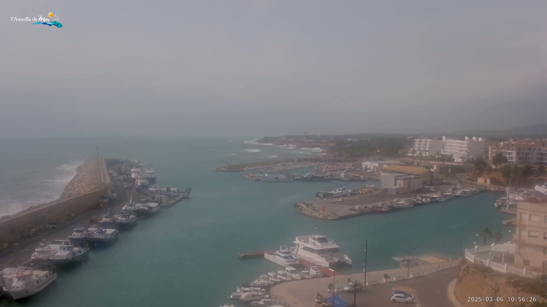 A hazy day reveals a harbor filled with numerous boats and ships, alongside a coastline featuring buildings and a wave-breaking structure.