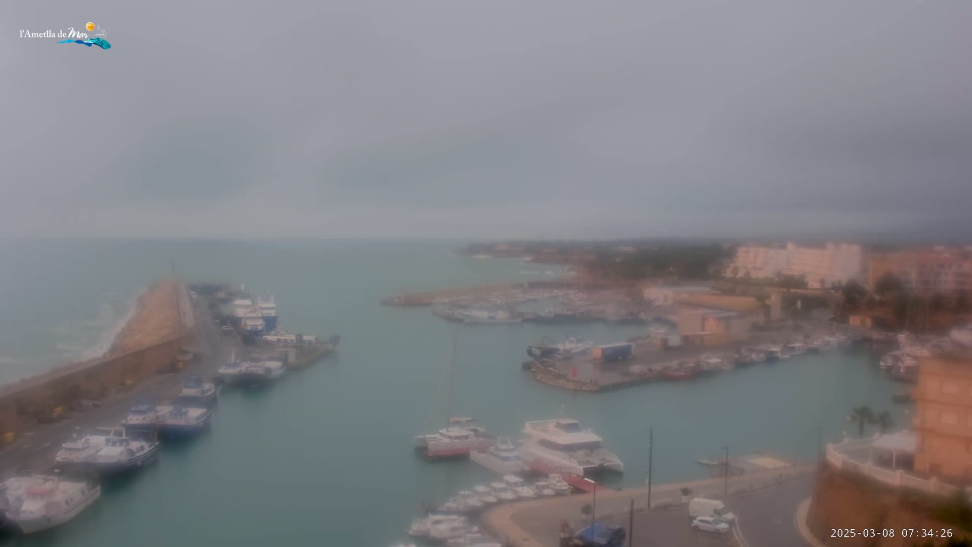 A hazy, overcast view shows a harbor filled with numerous boats and yachts, situated next to a town with buildings and some vegetation, under a gray sky.