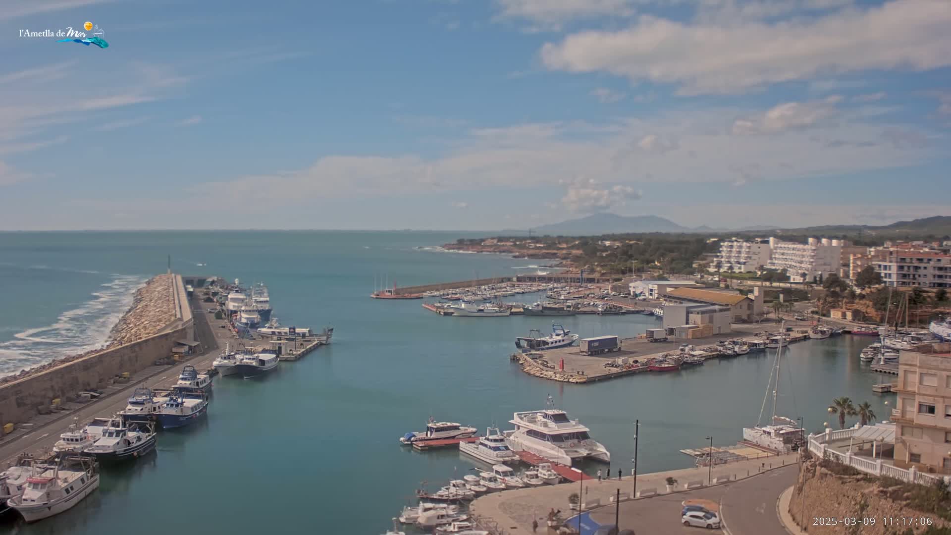 A harbor filled with numerous boats and yachts is situated next to a town under a partly cloudy sky, with a long stone breakwater protecting it from the ocean's waves.