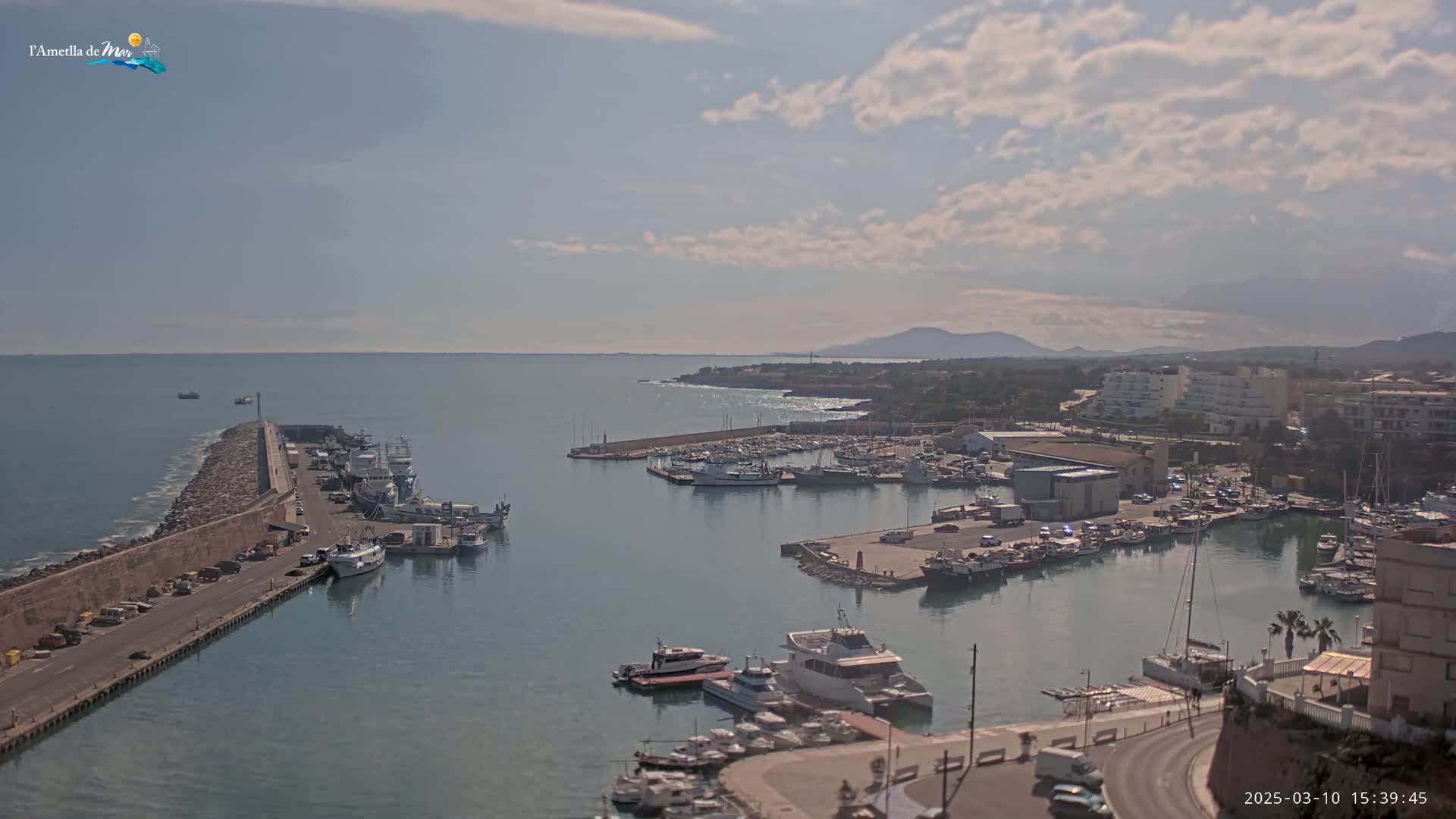 A partly cloudy day shows a harbor filled with boats, a stone breakwater, and a coastal town under a mostly sunny sky.