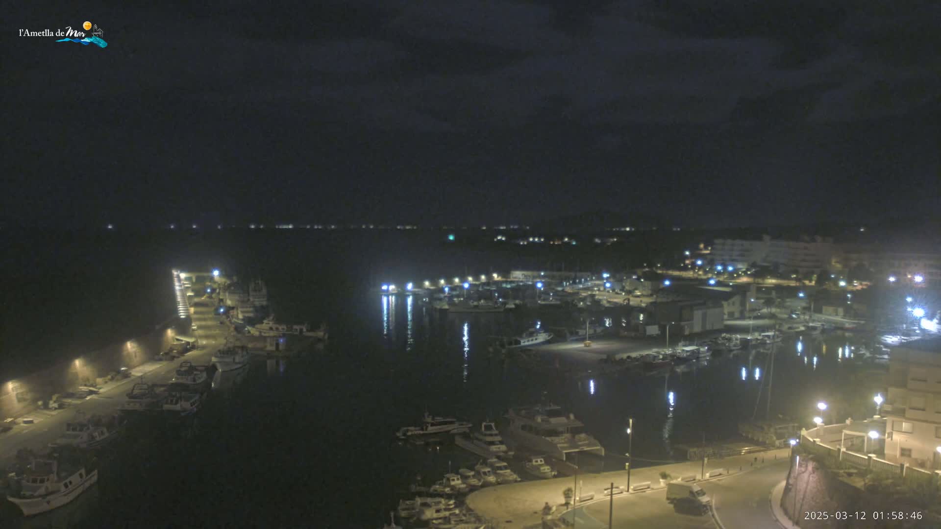 A nighttime, high-angle view of a harbor filled with boats, with surrounding buildings and streetlights illuminated under a dark, cloudy sky.