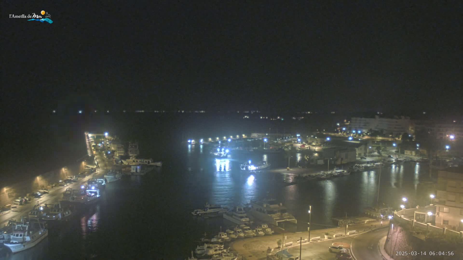 A nighttime view of a harbor filled with boats, illuminated by lights from buildings and street lamps along the waterfront.