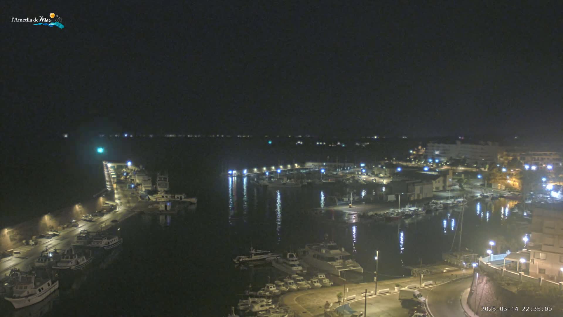 A nighttime aerial view of a harbor filled with boats, with the lights of buildings and streetlights reflecting on the calm water.