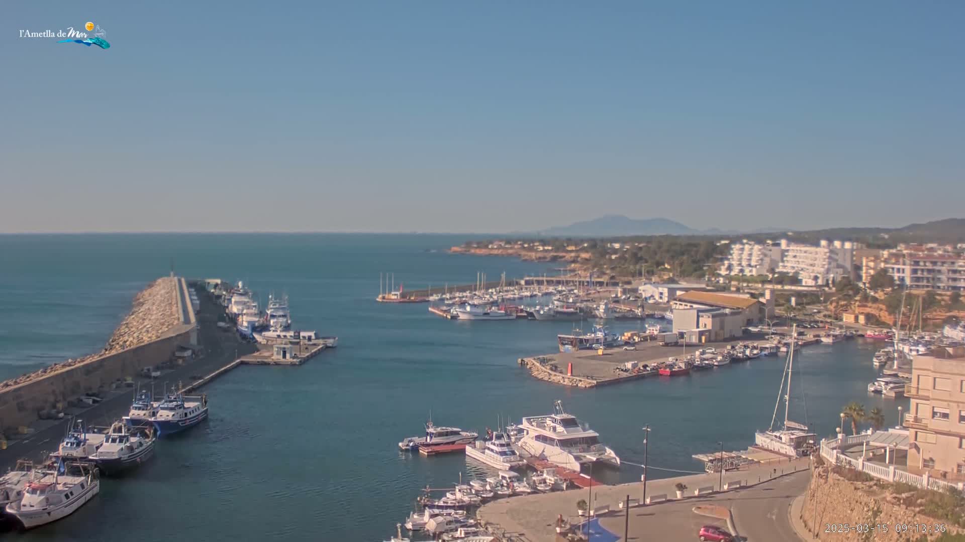A harbor filled with numerous boats and yachts is shown on a sunny day, with calm water and a distant mountain range visible on the horizon.