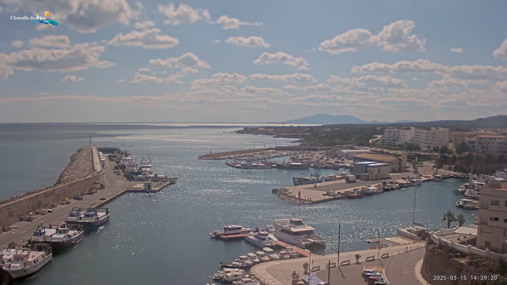 A harbor filled with boats is seen under a partly cloudy sky, with a distant coastline and mountains visible in the background.