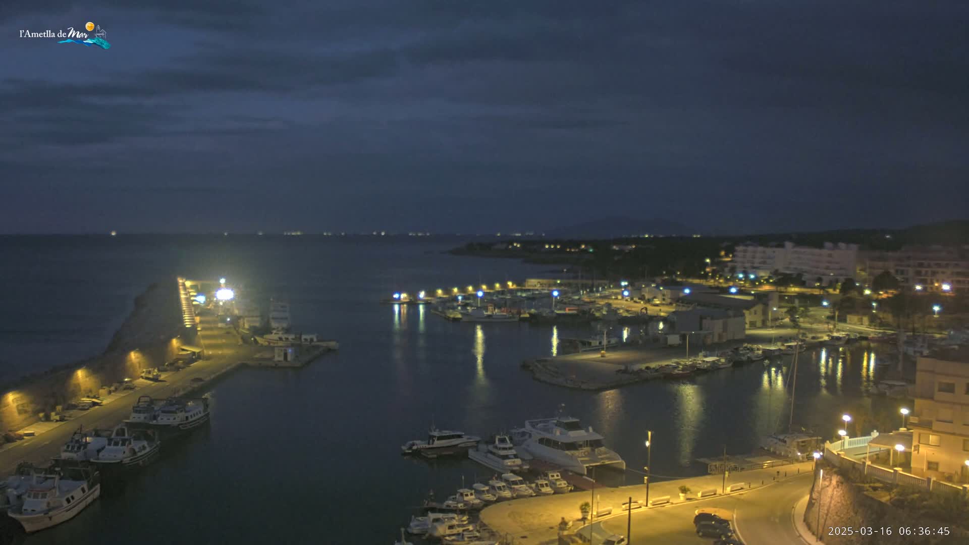 A nighttime view of a harbor filled with boats, lit by streetlights and building lights under a dark, cloudy sky.