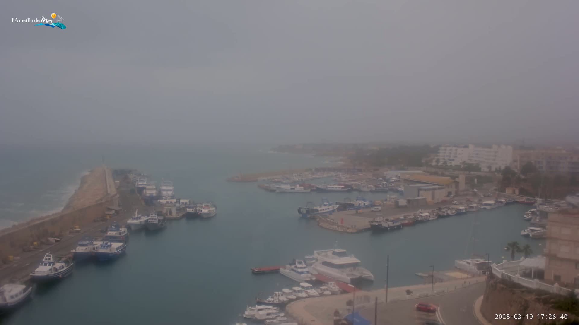 A hazy harbor filled with numerous boats is seen from an elevated perspective, with a stone breakwater extending into a somewhat choppy sea.