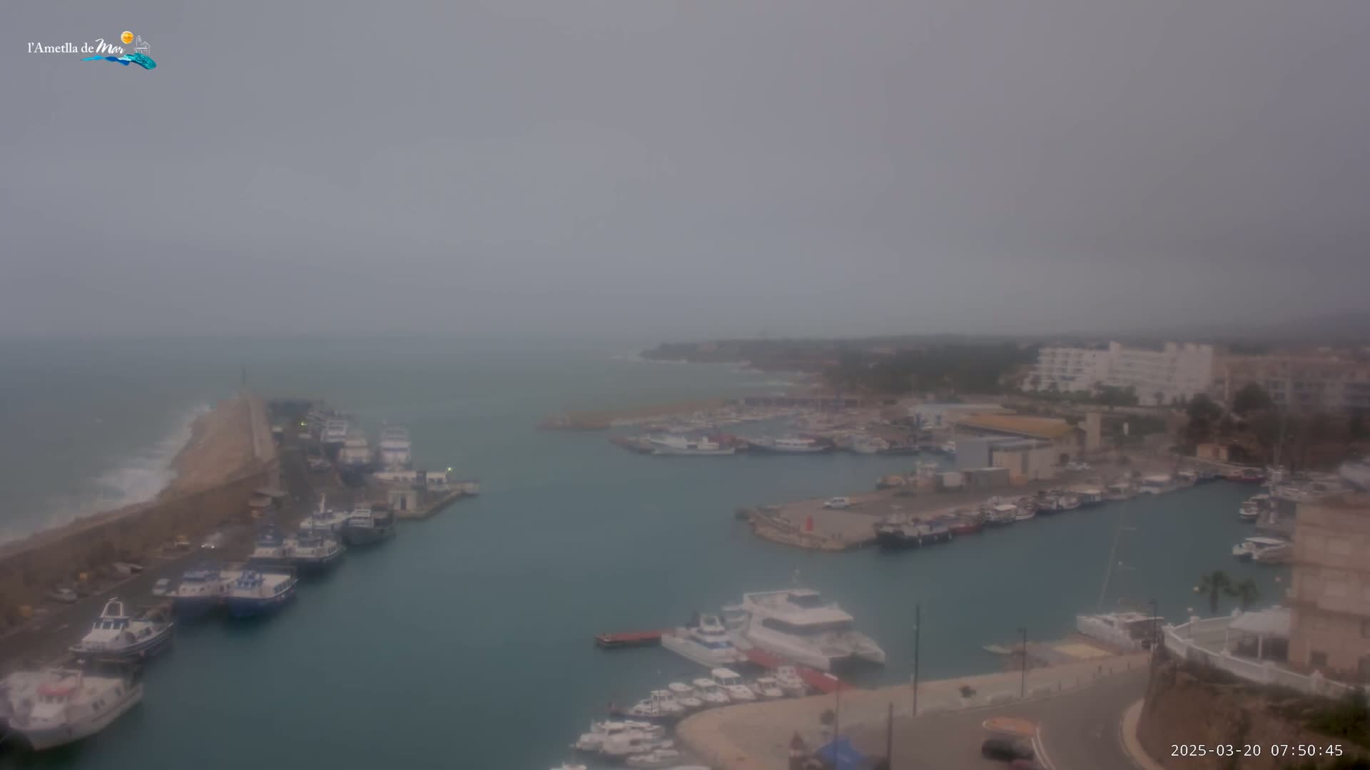 A harbor filled with numerous boats is shown under an overcast sky, with waves crashing against a seawall.