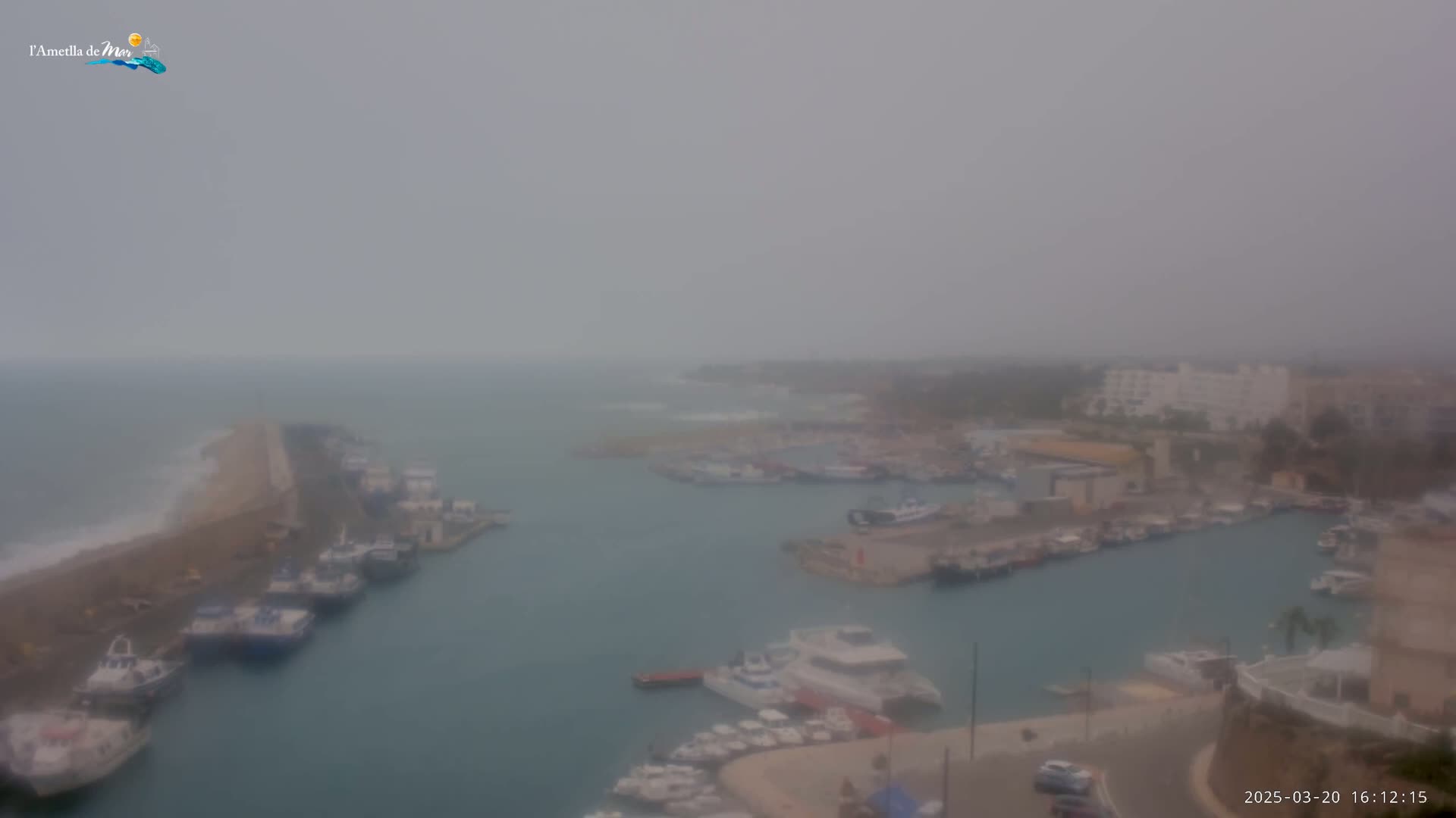 A hazy, overcast day reveals a harbor filled with numerous boats and ships, nestled between a rocky shoreline and a coastal town.