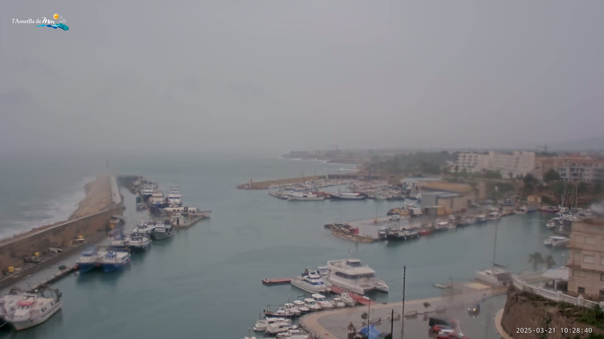 A harbor filled with numerous boats is seen on an overcast day, with waves crashing against a breakwater in the distance.