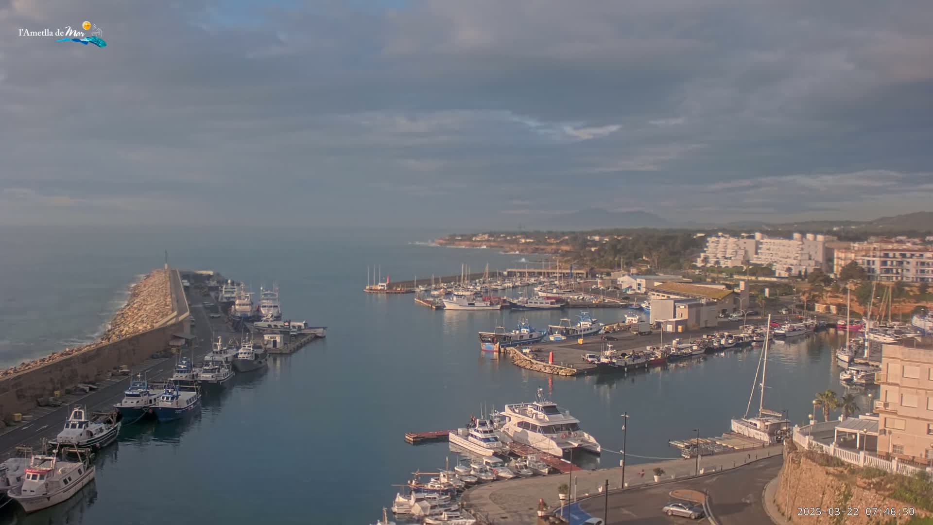 A harbor filled with numerous boats and yachts is situated near a coastal town under a mostly cloudy sky.