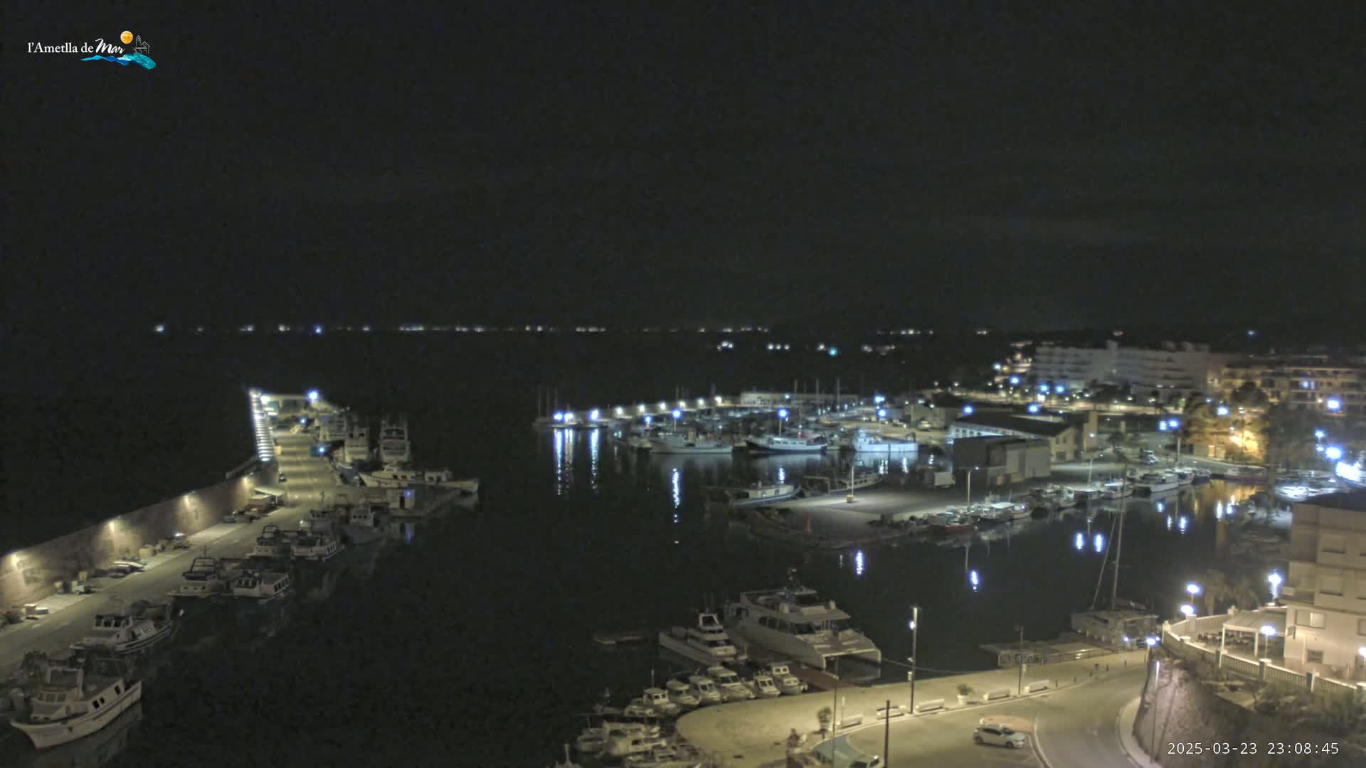 A nighttime, aerial view of a harbor filled with boats, illuminated by lights, with a town in the background under a clear sky.