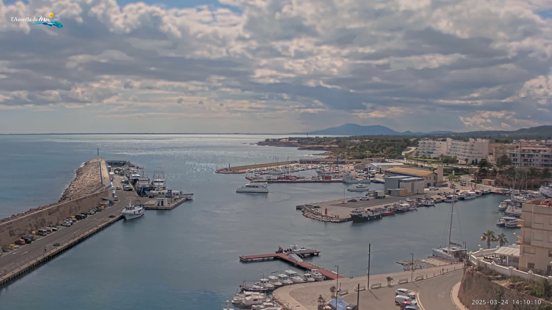 A partly cloudy day overlooks a harbor filled with boats and buildings, with a calm sea extending to the horizon and mountains visible in the distance.