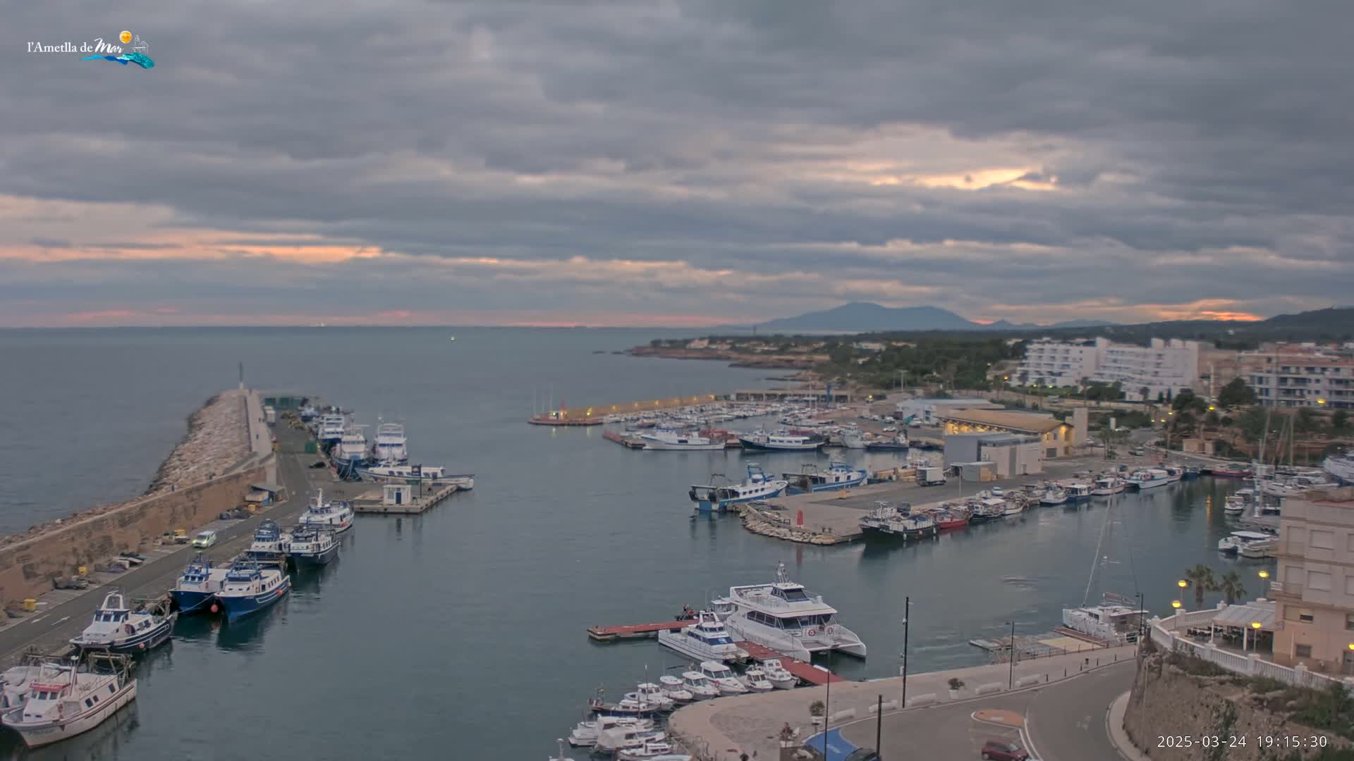 A harbor filled with numerous boats and yachts is nestled along a coastline under a cloudy sunset sky.