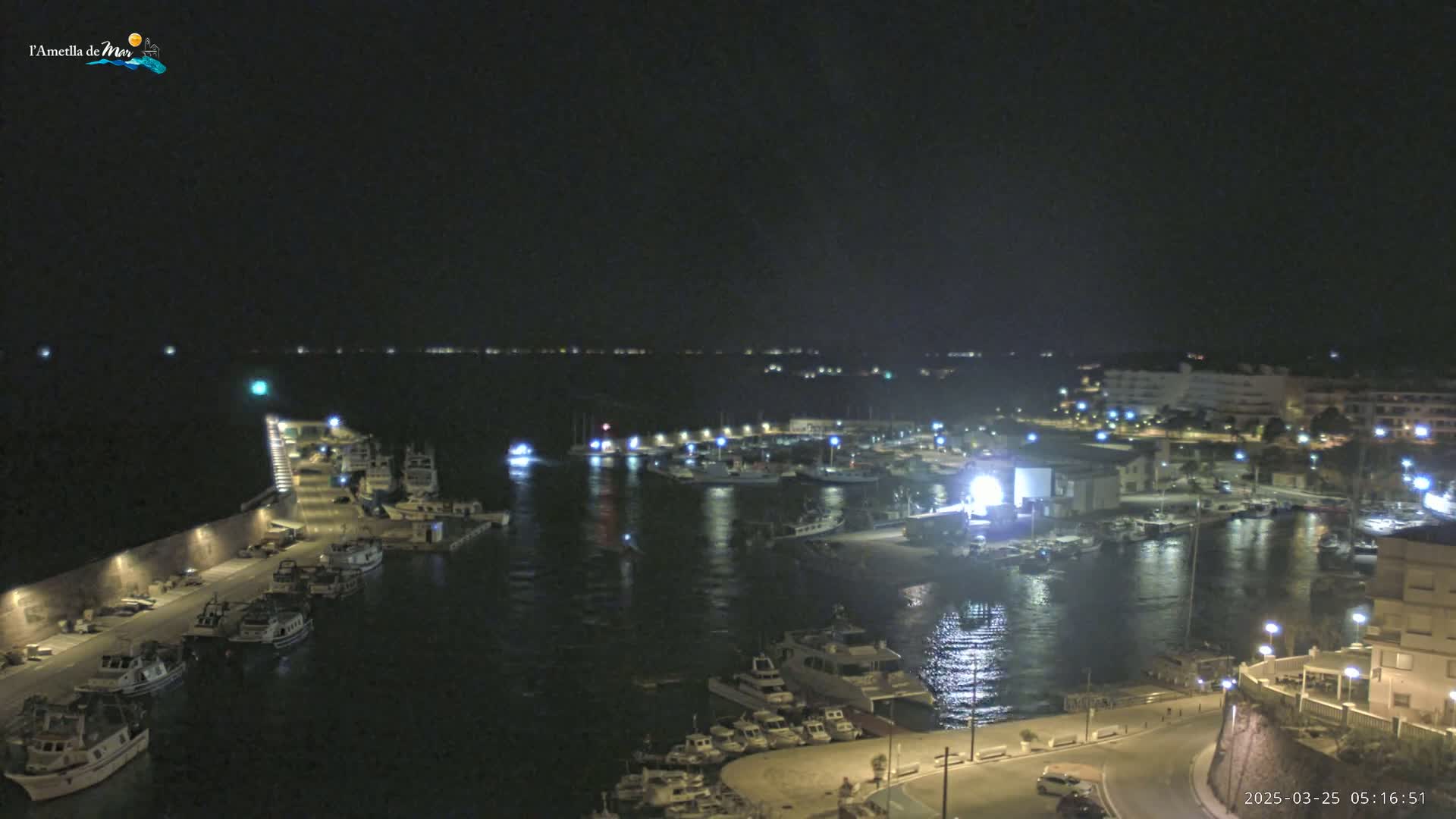 A nighttime, aerial view of a harbor filled with boats, with calm water reflecting the lights of the harbor and surrounding buildings.