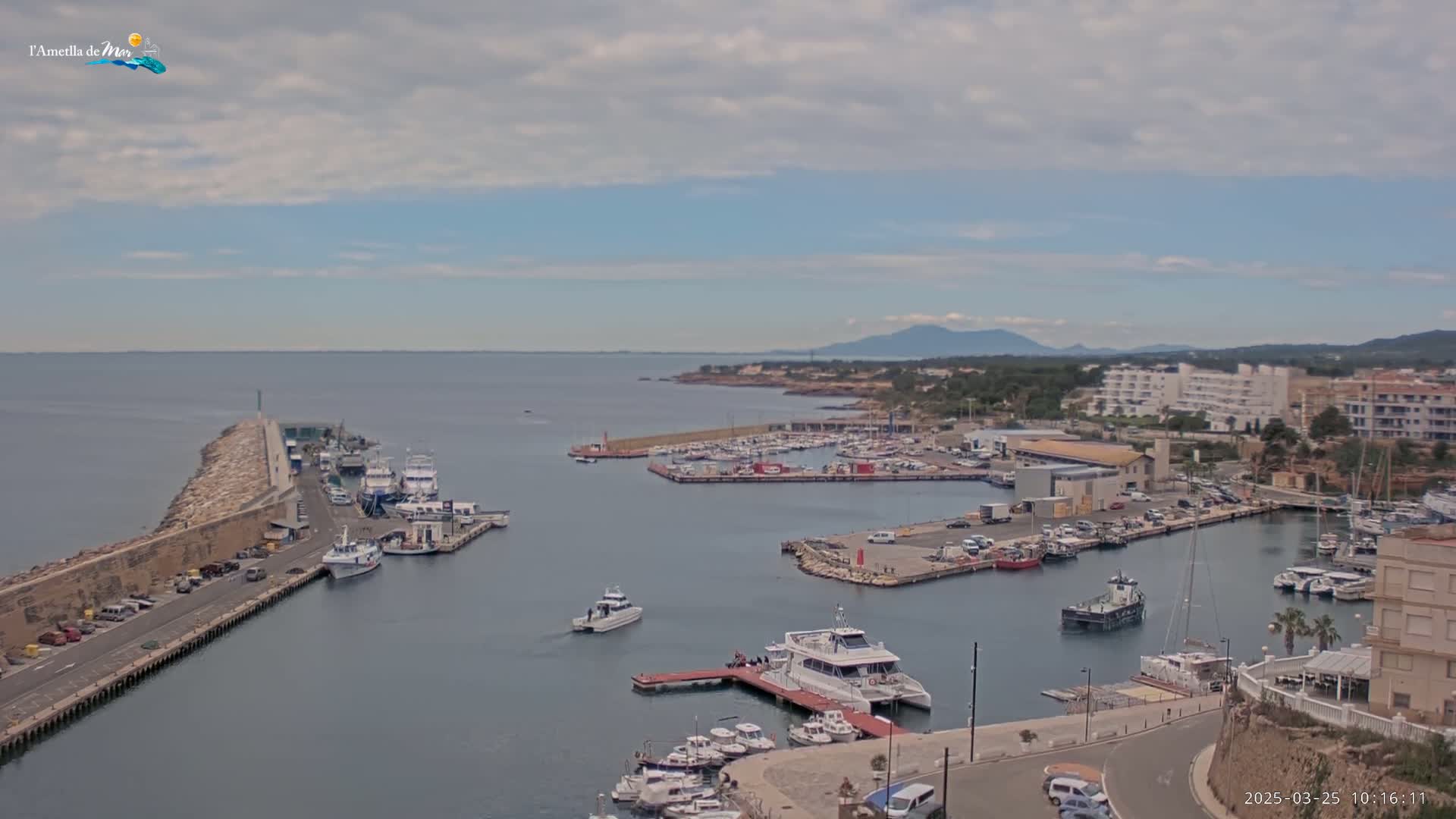 A harbor filled with boats is shown under a mostly cloudy sky, with a town and distant mountains visible in the background.