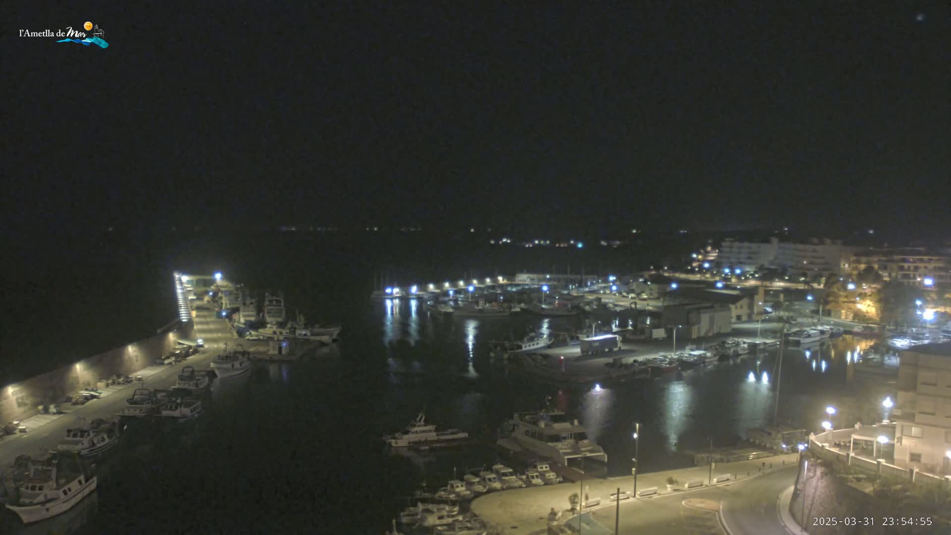 A nighttime view of a harbor filled with boats, illuminated by lights along the docks and buildings on the shore under a clear, dark sky.