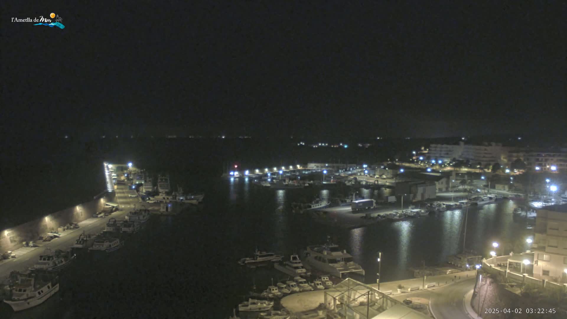 A nighttime, aerial view of a harbor filled with boats, docked along a waterfront with buildings and lights illuminating the scene.